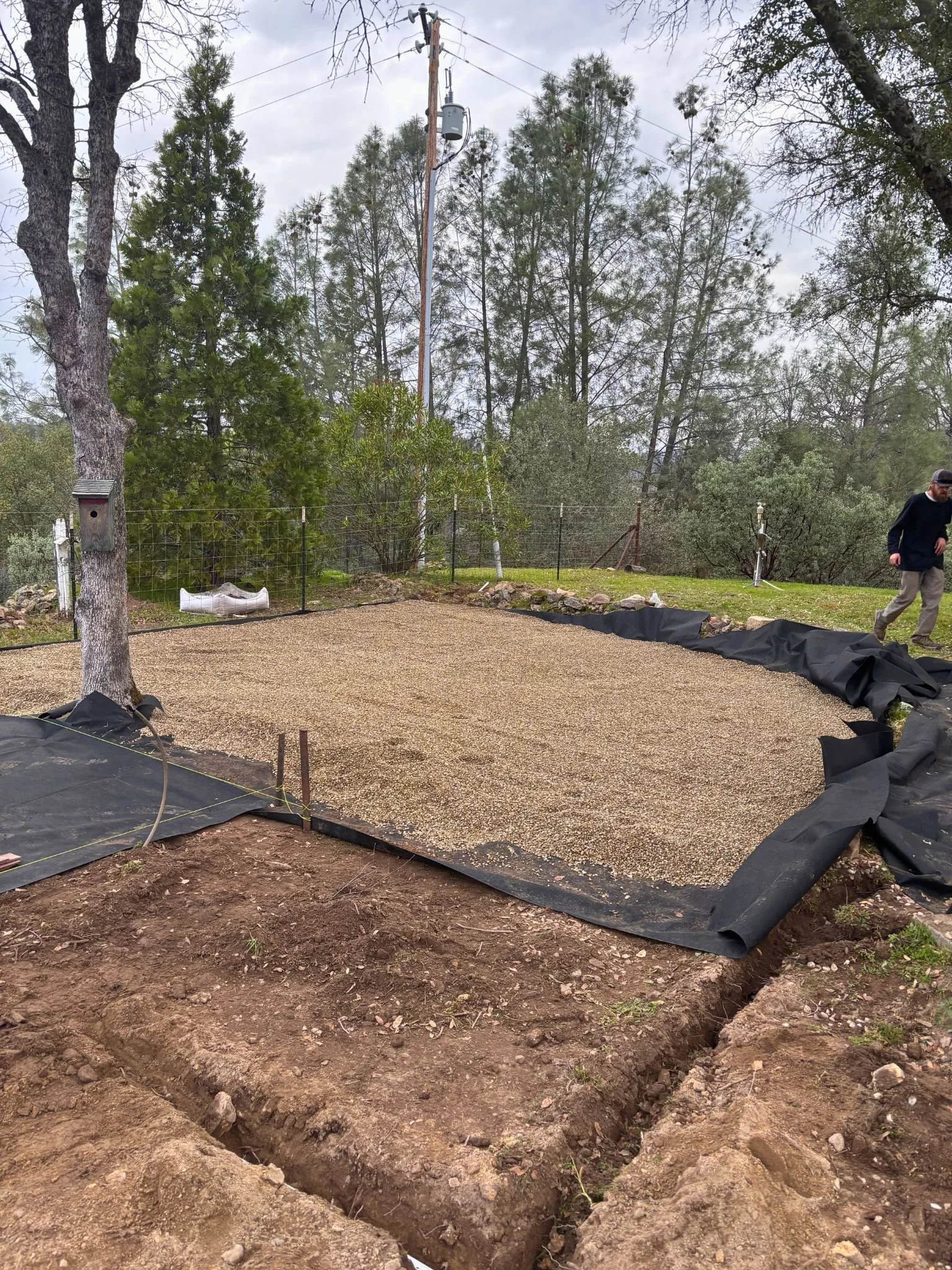 A large rectangular area of soil with a trench dug around its perimeter, black plastic sheeting along the edges, and a gravel surface in the center. In the background, there are trees, a fence, and a person walking on the right side.