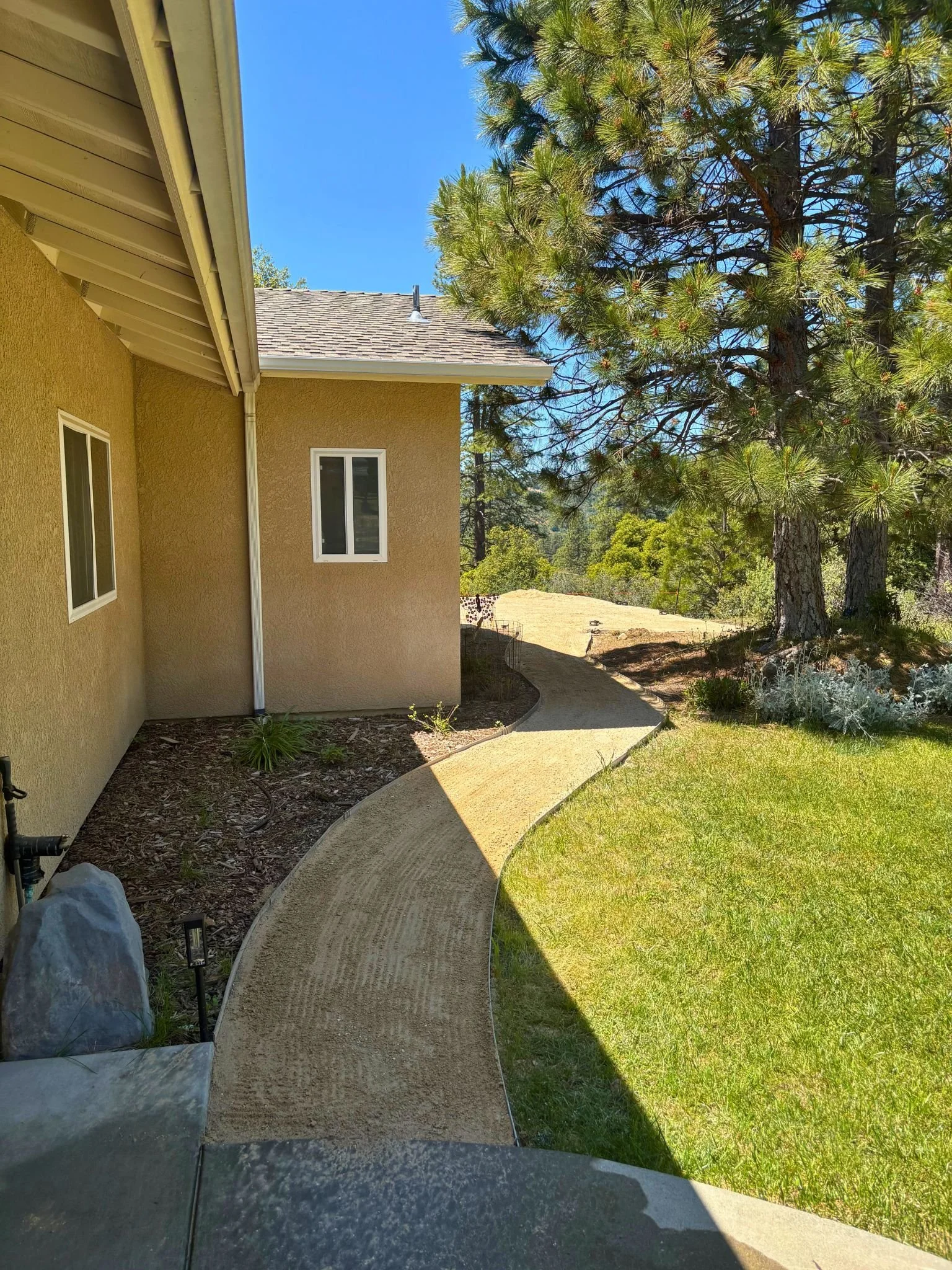 A pathway curves along the side of a house with yellowish stucco walls, leading to a garden with tall pine trees and green grass. The house has a window and an overhanging roof with visible eaves.
