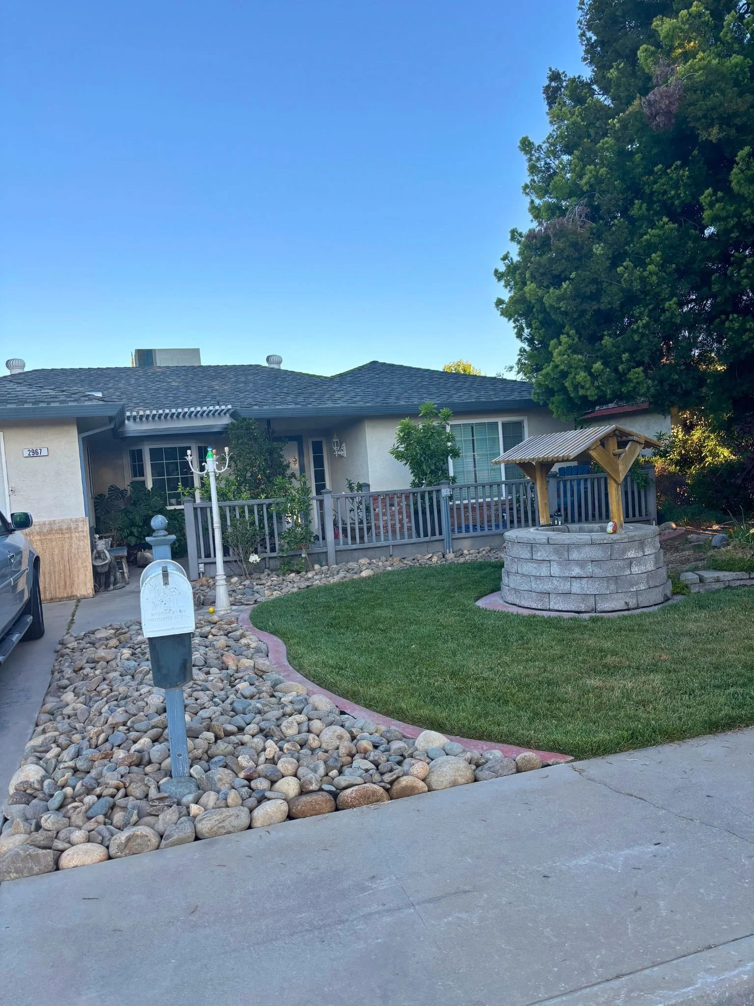 A house with a gray roof, white walls, and a front yard with a stone well, a small garden, and a wooden fence. There is a large tree on the right and some decorative rocks along the walkway.