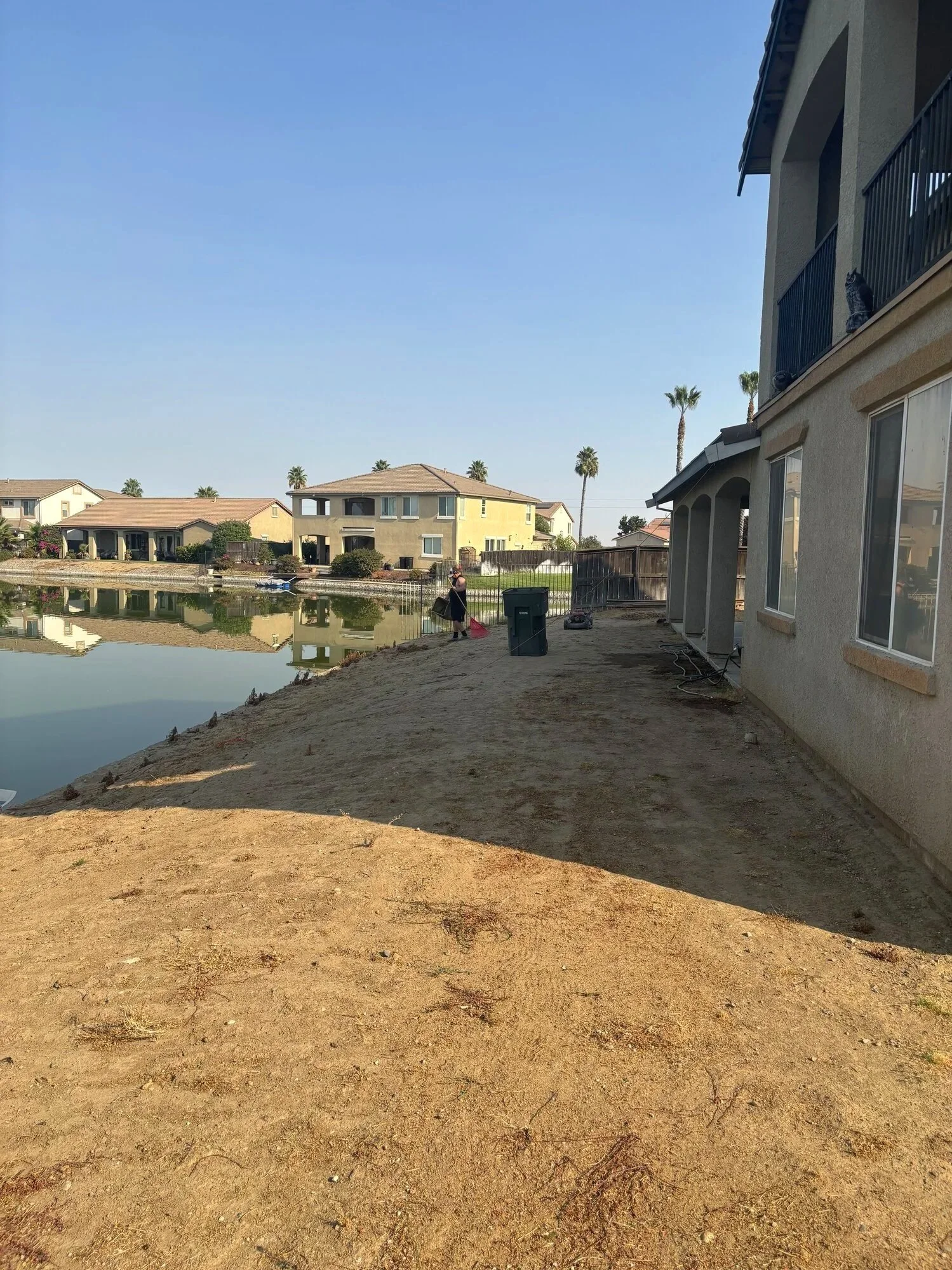 A backyard with a dirt area, next to a house with a fenced patio, and across a body of water with residential houses and palm trees in the background.