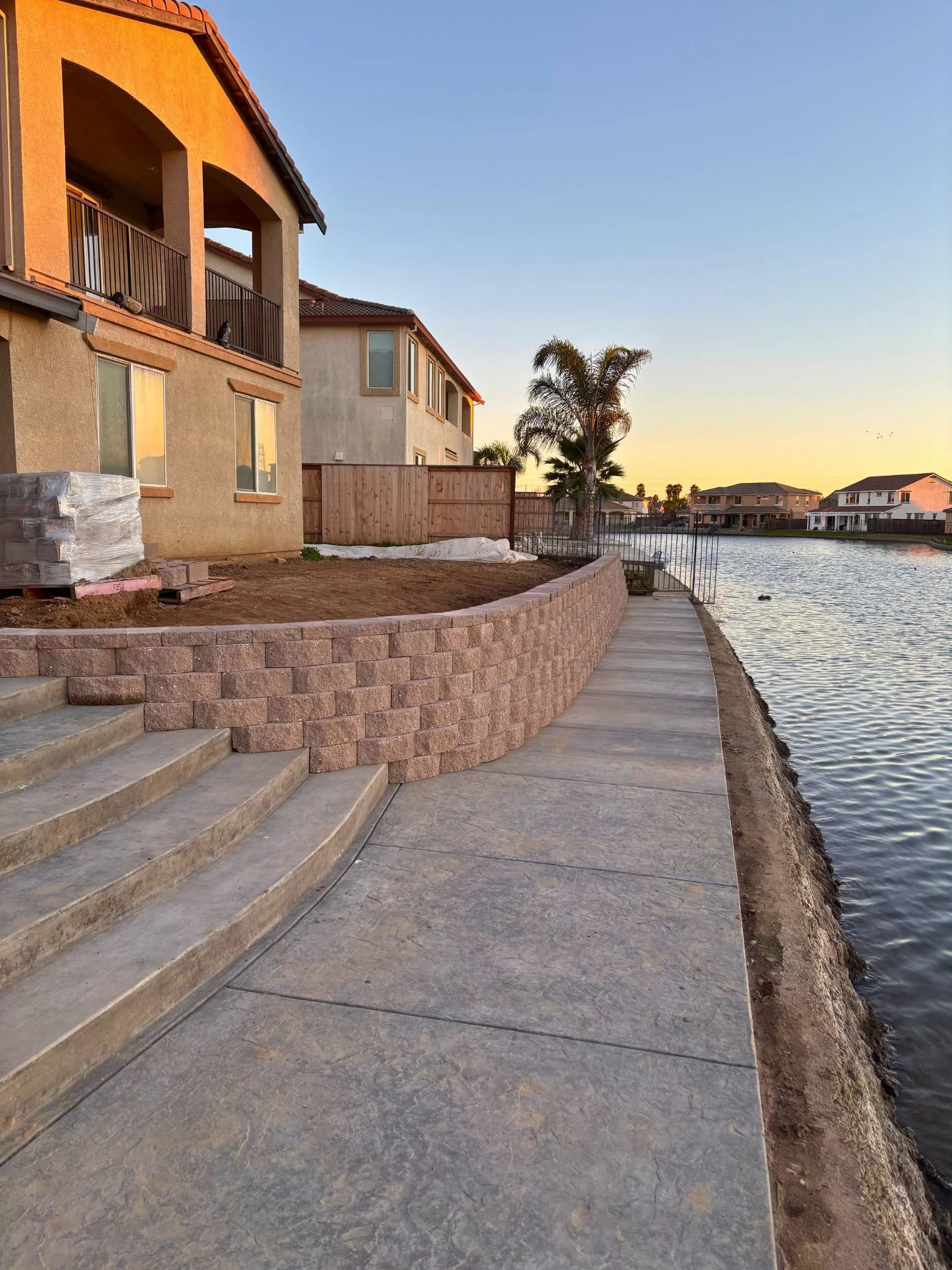 A residential backyard by a canal at sunset, featuring a multi-story house, a palm tree, and a walking path with steps.