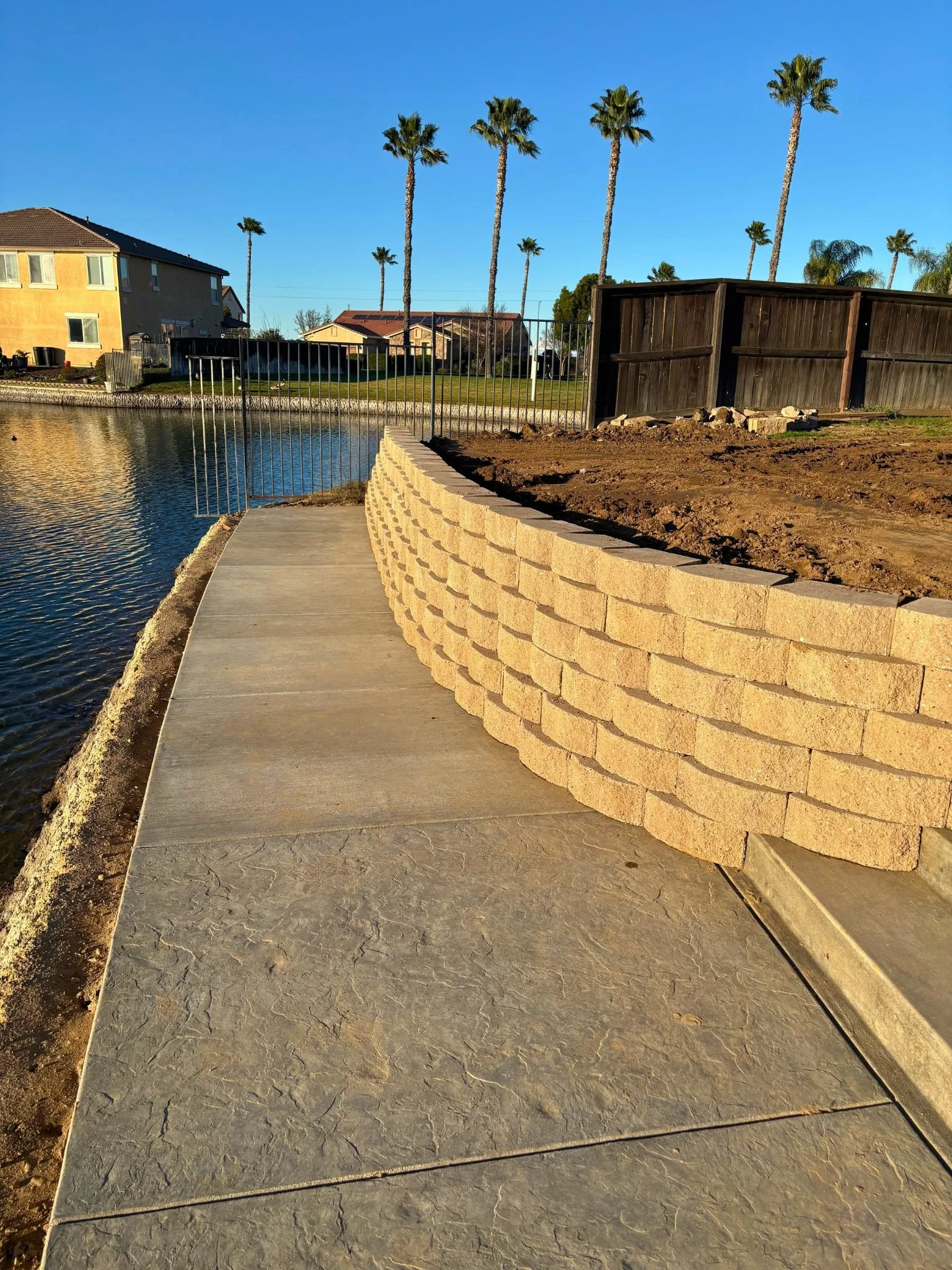 A sidewalk along a body of water with a stone retaining wall on one side and a wooden fence on the other. Houses and tall palm trees are in the background under a clear blue sky.