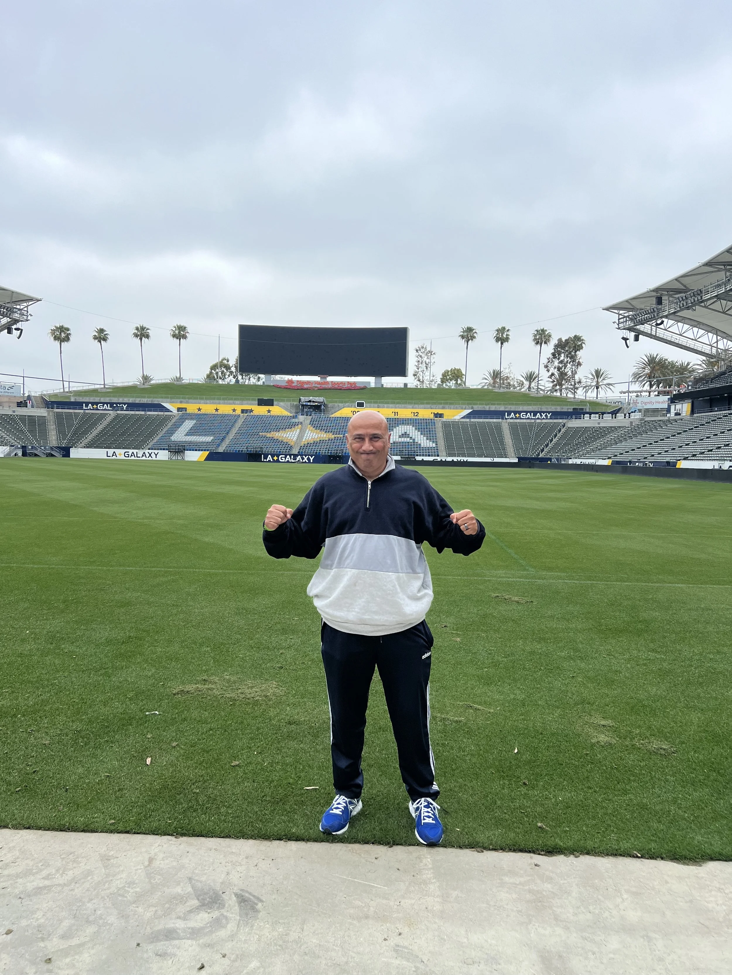 Jake Cancik, or Agop Cancik standing on a green football field with arms raised, wearing a black and white jacket, black pants, and blue shoes, in a stadium with empty seats and a large digital screen in the background