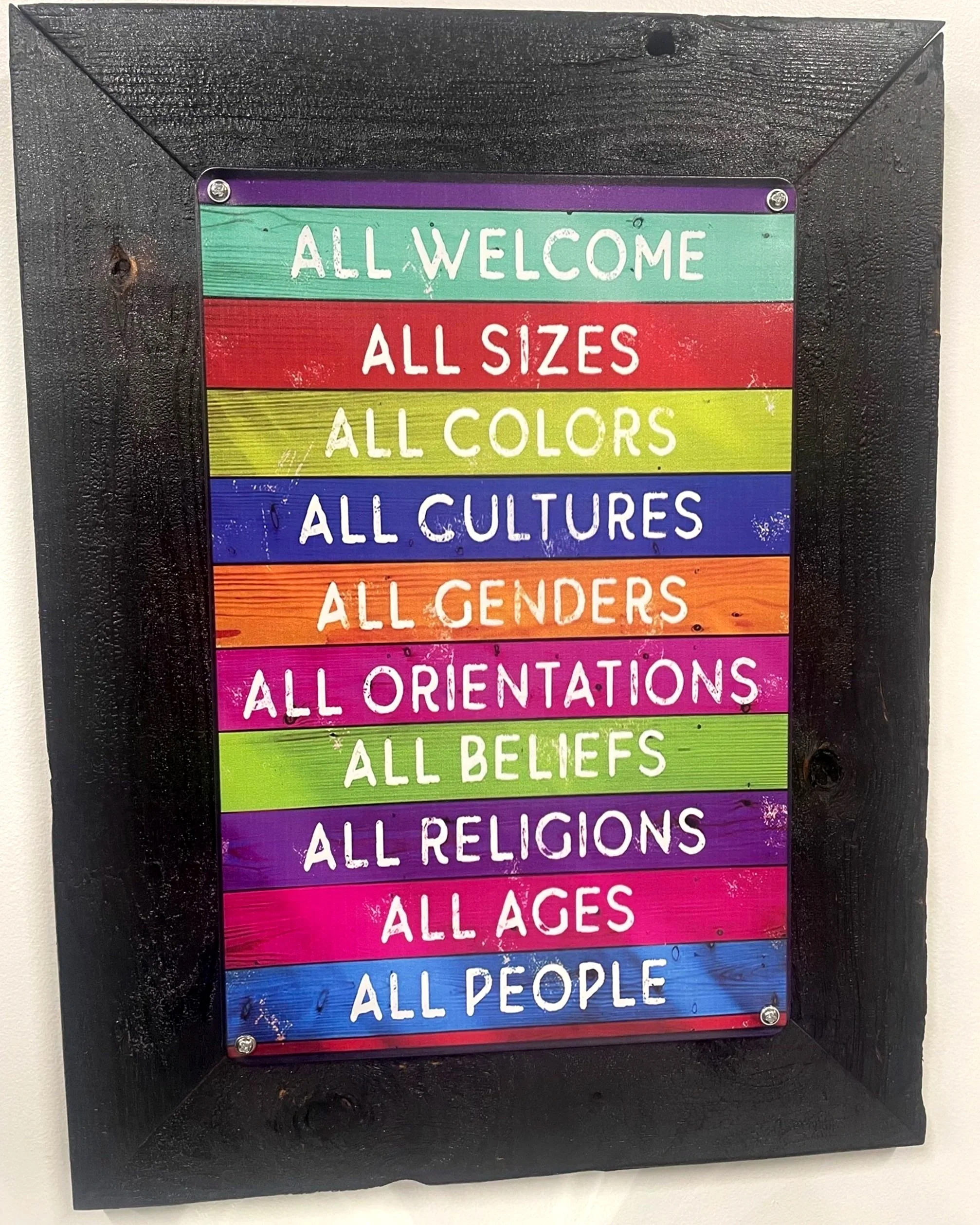 Colorful sign with rainbow stripes and white text that reads 'All Welcome, All Sizes, All Colors, All Cultures, All Genders, All Orientations, All Beliefs, All Religions, All Ages, All People' framed by black wood.