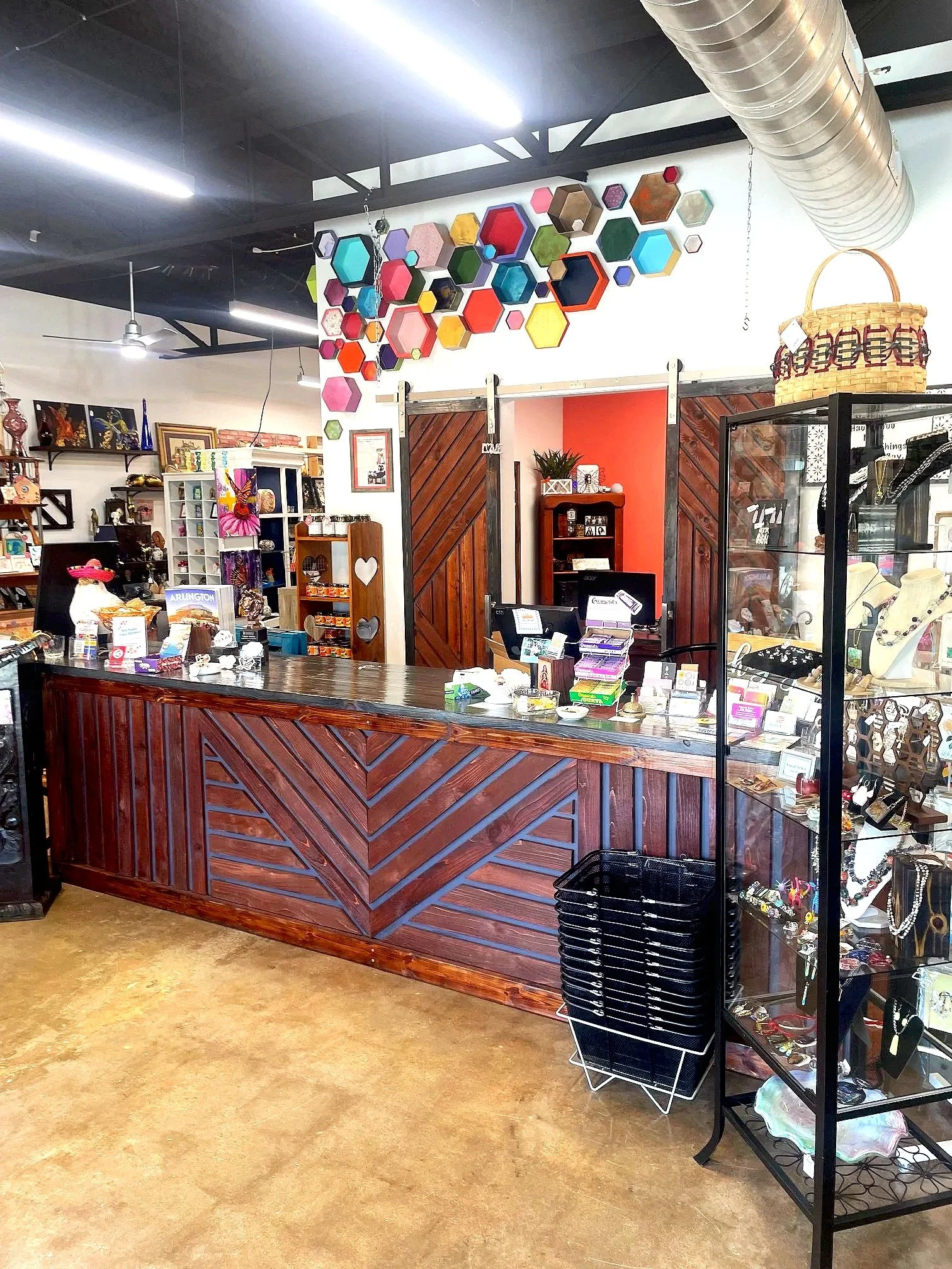 The Mosaic Market front counter, colorful hexagon-shaped wall art, display shelves with jewelry and decor, and a barn-style door with a red accent wall in the background.