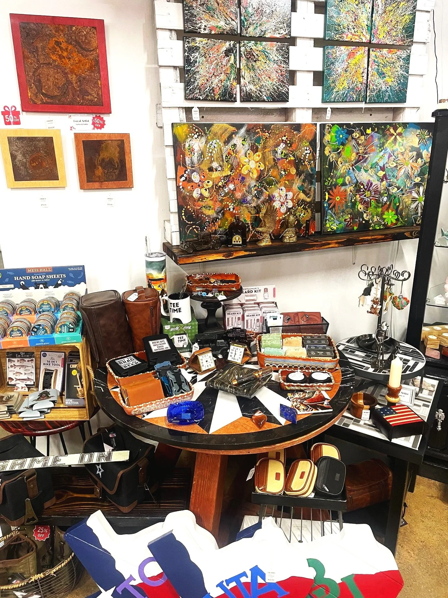 Various abstract paintings hanging on the wall and on display shelves inside The Mosaic Market art and gift store, with a round table in the center filled with small decorative items and accessories.