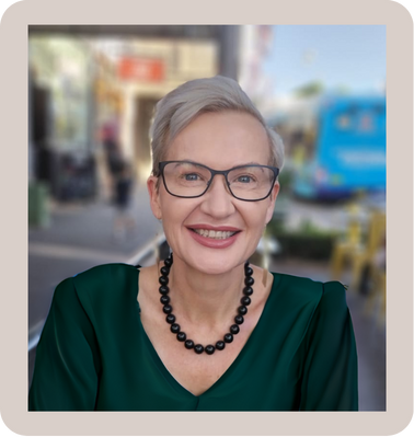 Smiling senior woman with short gray hair and glasses, wearing a green top and black beaded necklace, outdoors with a blurred city street background.