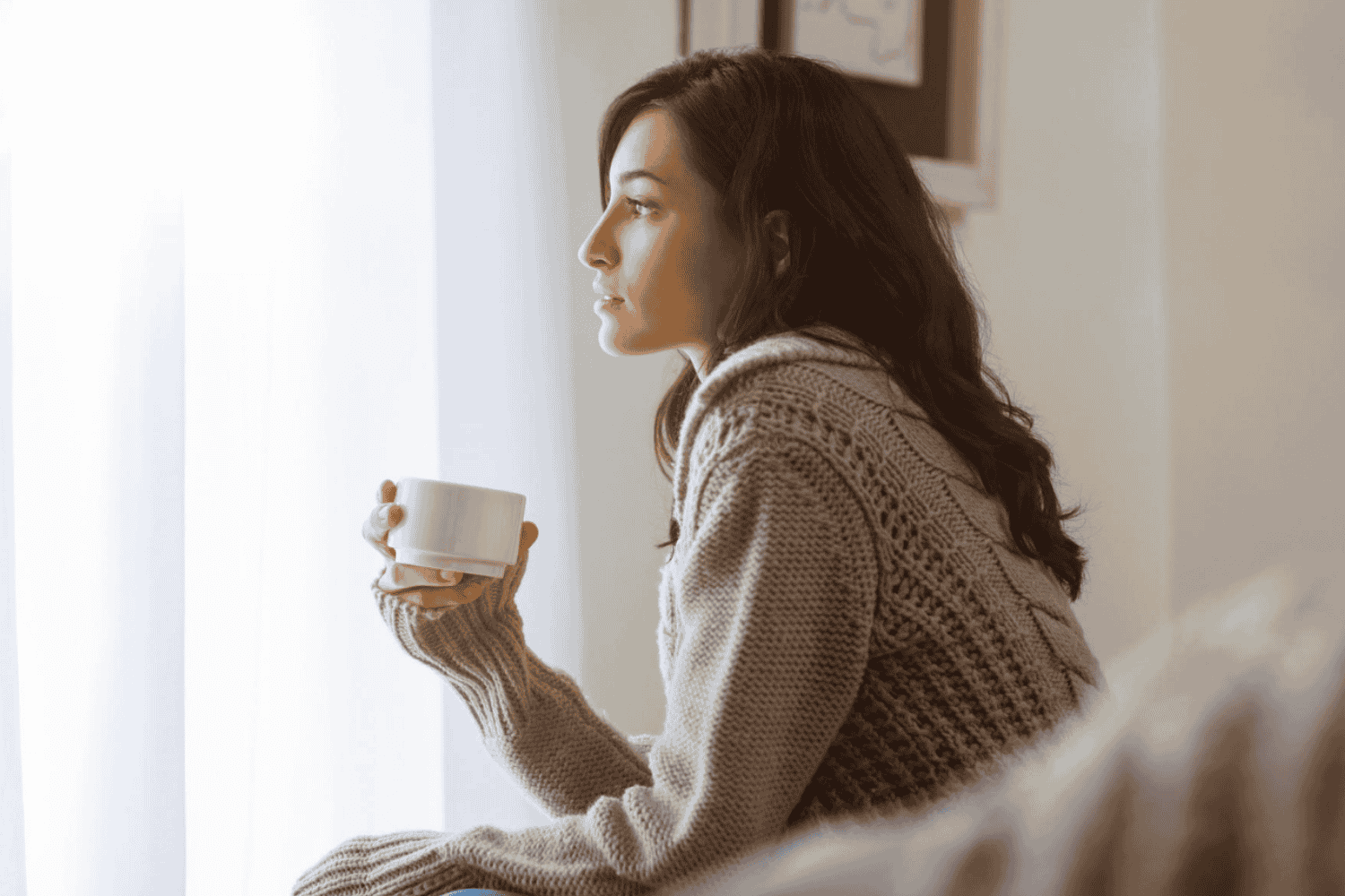 Woman sitting by window holding a white mug, wearing a cozy knitted sweater, looking contemplative.