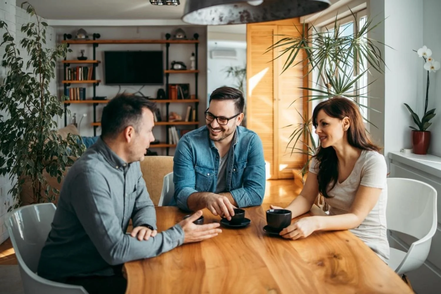 Three people sitting around a wooden table in a cozy living room, smiling and having a conversation while holding coffee cups.