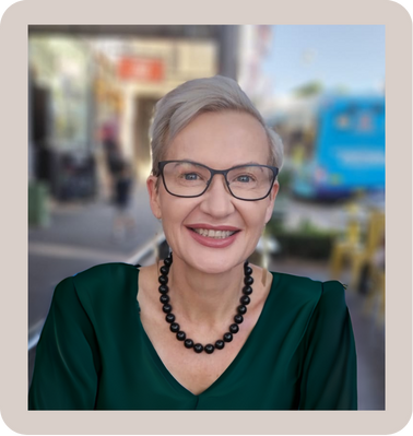 ngahina richards Smiling with short gray hair and glasses, wearing a green blouse and a black beaded necklace, outdoors in an urban setting.
