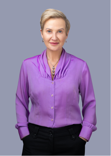 A woman with short blonde hair wearing a purple blouse and black pants, standing against a light blue background.