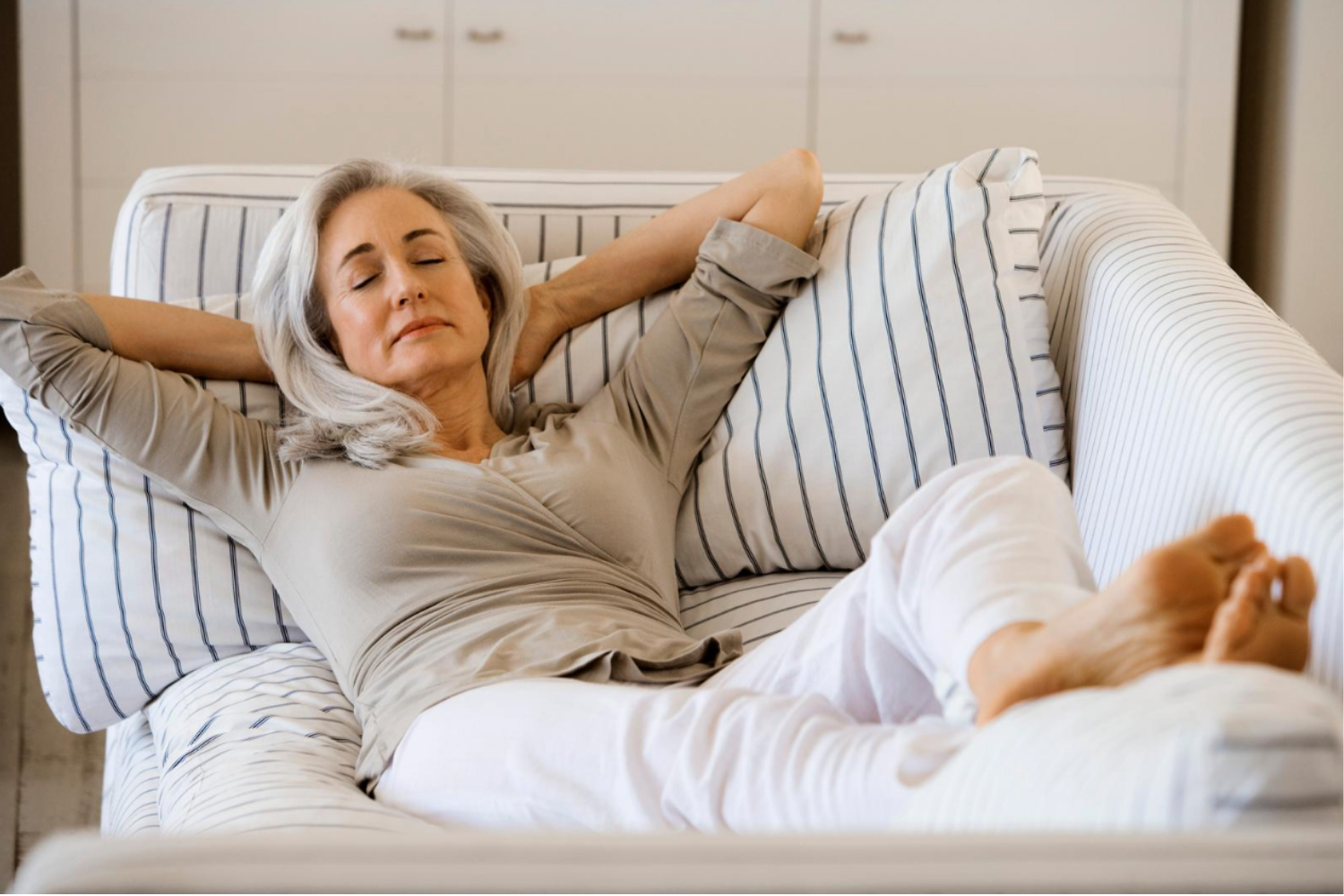 An older woman with gray hair is relaxing on a striped couch with her eyes closed and arms resting behind her head.