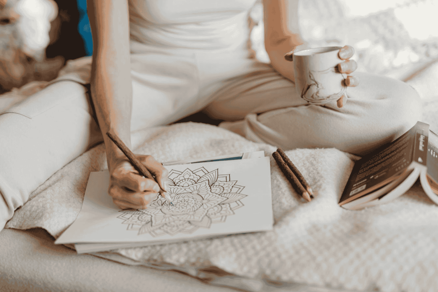Person sitting on a bed, coloring a detailed mandala design with colored pencils, holding a coffee mug in their right hand, with a book and additional coloring supplies nearby.