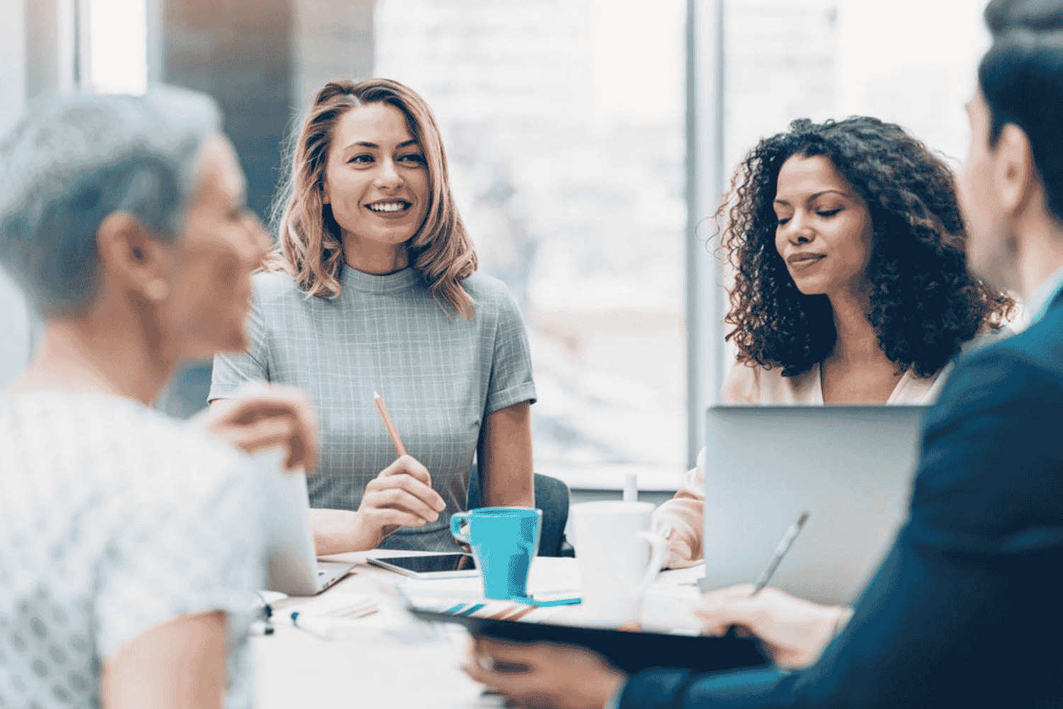 Four women engaged in a conversation around a table in a modern office, one woman smiling and holding a pen, two women with laptops, one woman appears to be speaking.