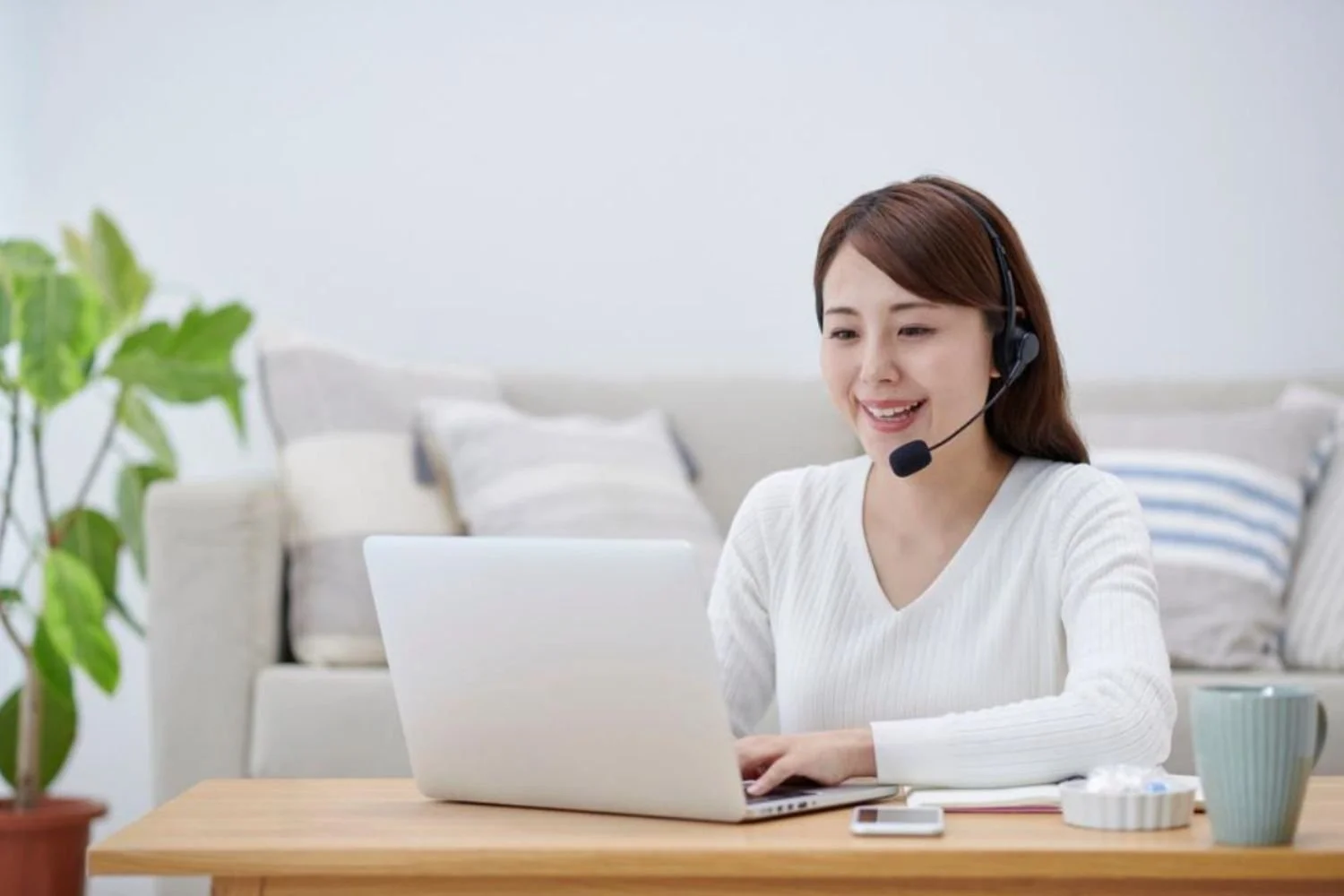 Woman wearing a headset working on a laptop, sitting at a wooden table with a potted plant nearby, in a cozy living room.