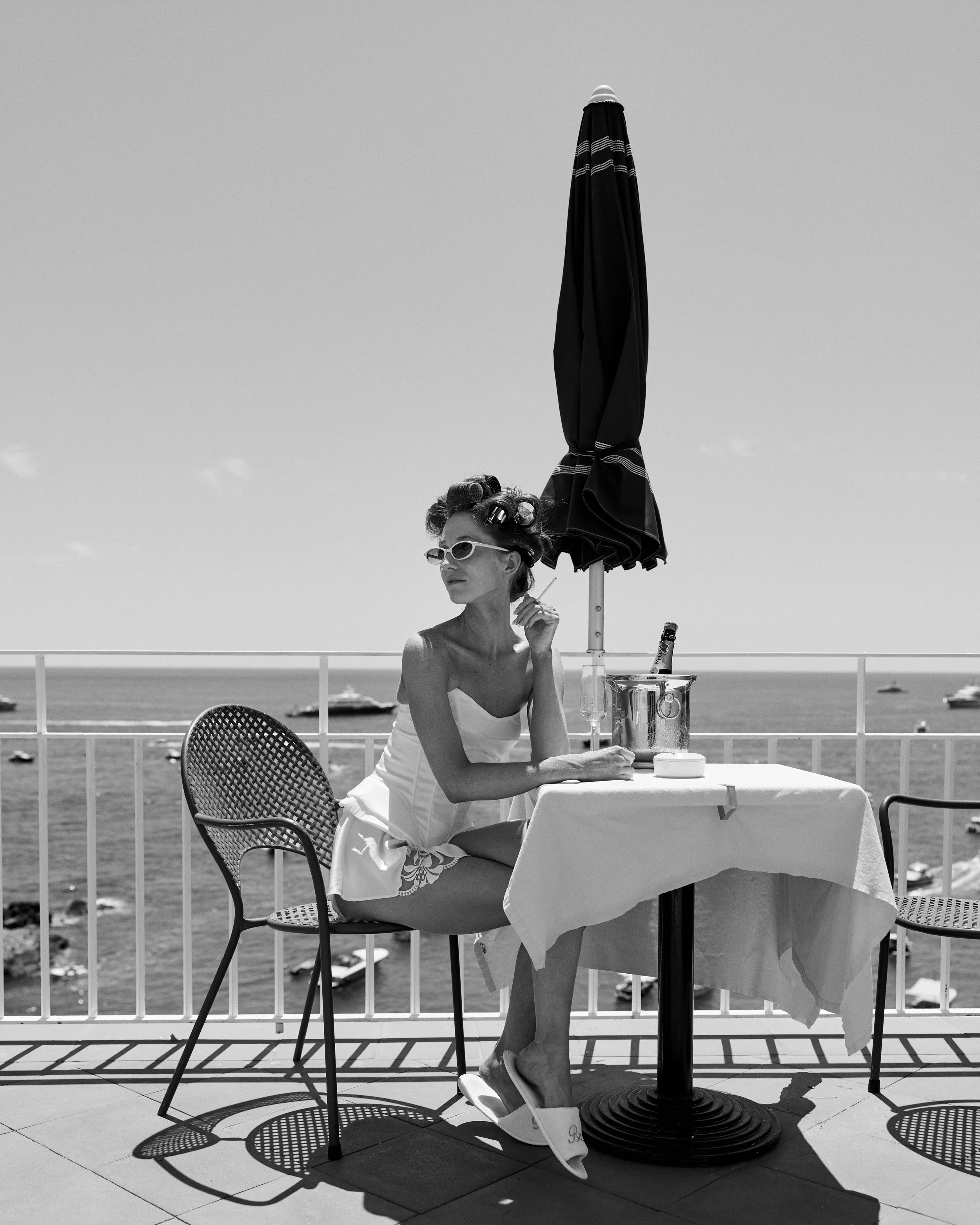 A woman with curlers in her hair and wearing sunglasses, sitting at an outdoor table with a tablecloth, on a balcony overlooking the ocean, with boats in the water and an umbrella in the background, in black and white.