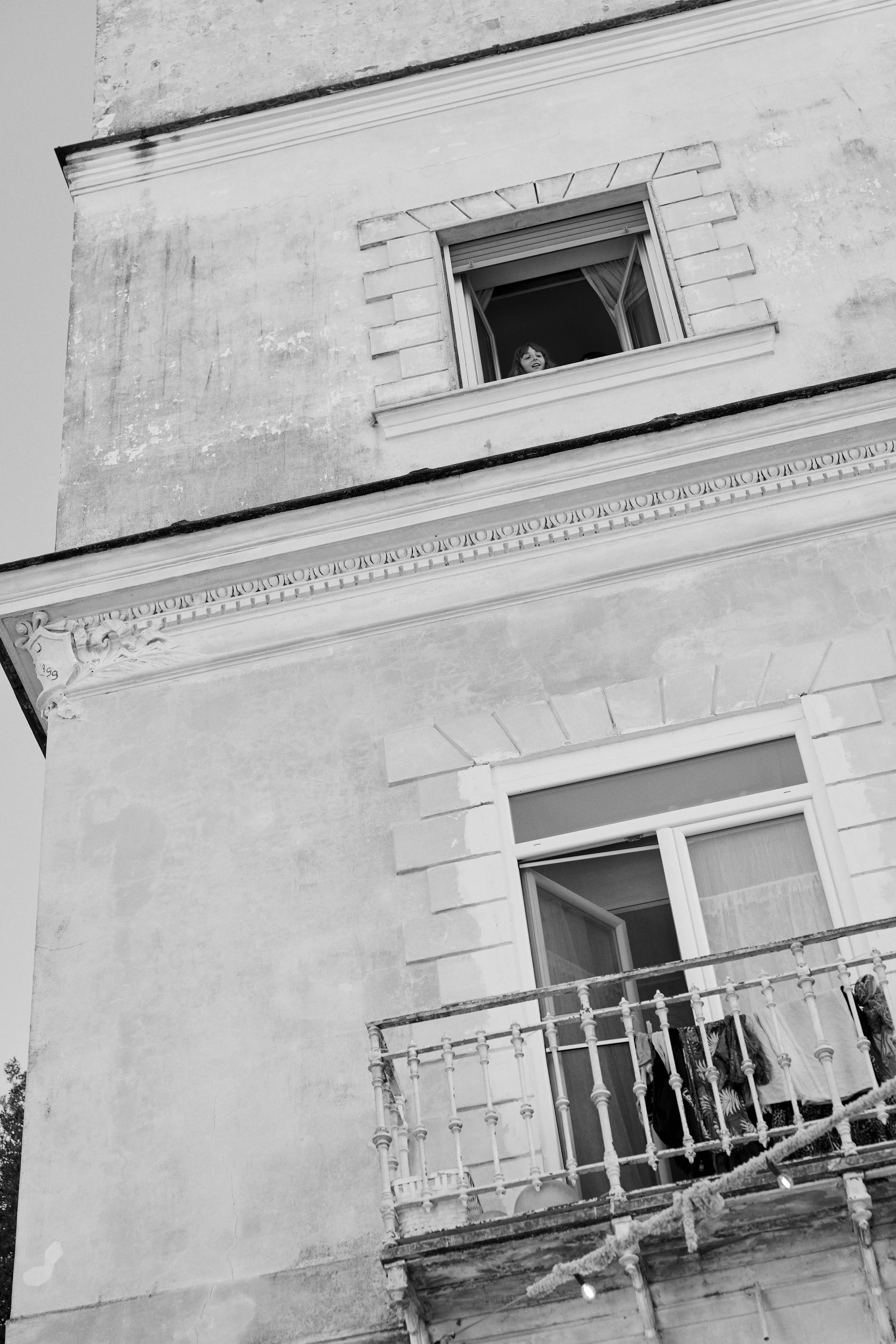 Black and white photo of the exterior of a weathered building with two windows, one on the lower level with an open door and a balcony with folded clothes, and one on the upper level with a child looking out.