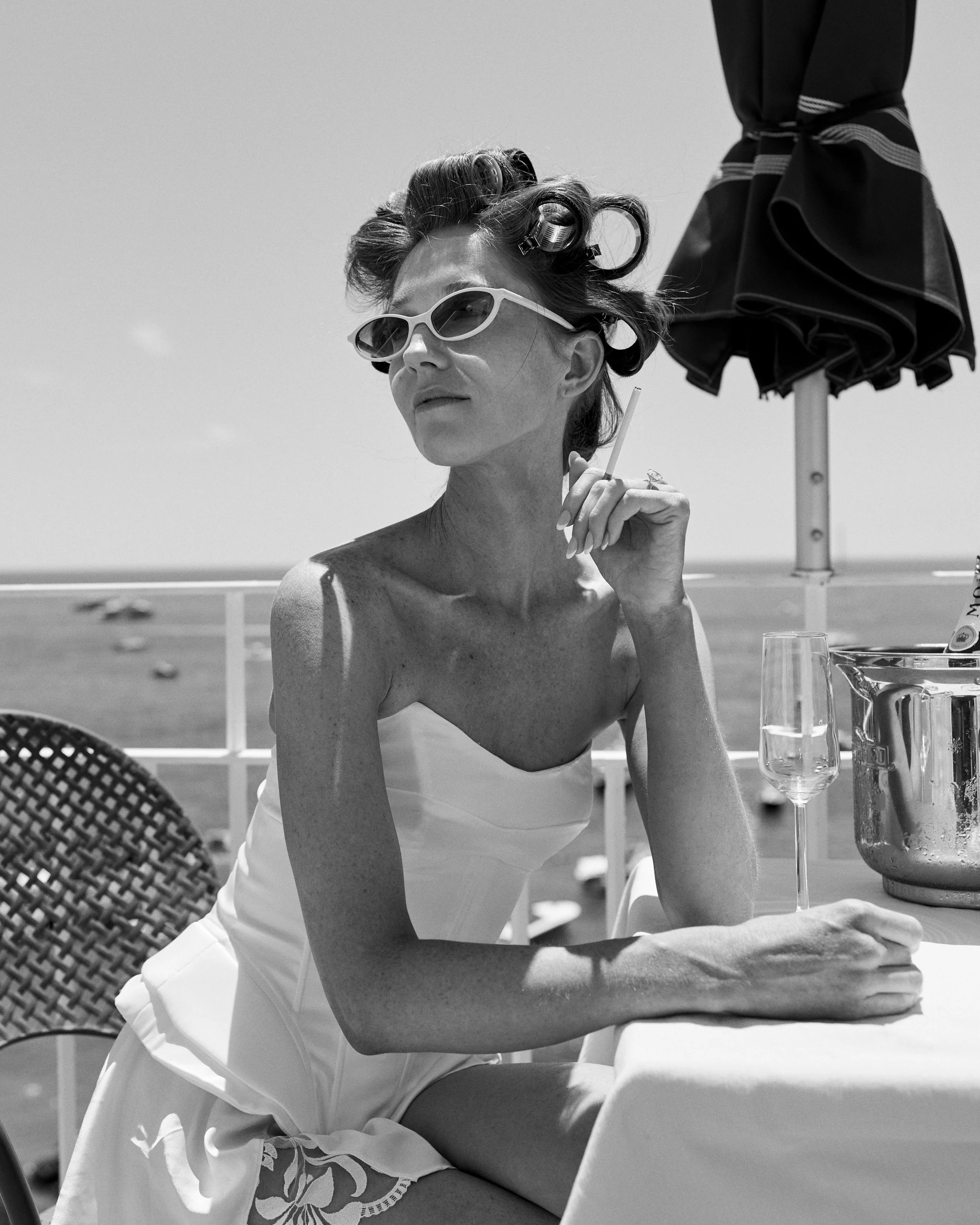 A woman in vintage style, wearing sunglasses and a strapless dress, sits at an outdoor table with a glass of champagne and an ice bucket nearby, with a beach and ocean in the background.