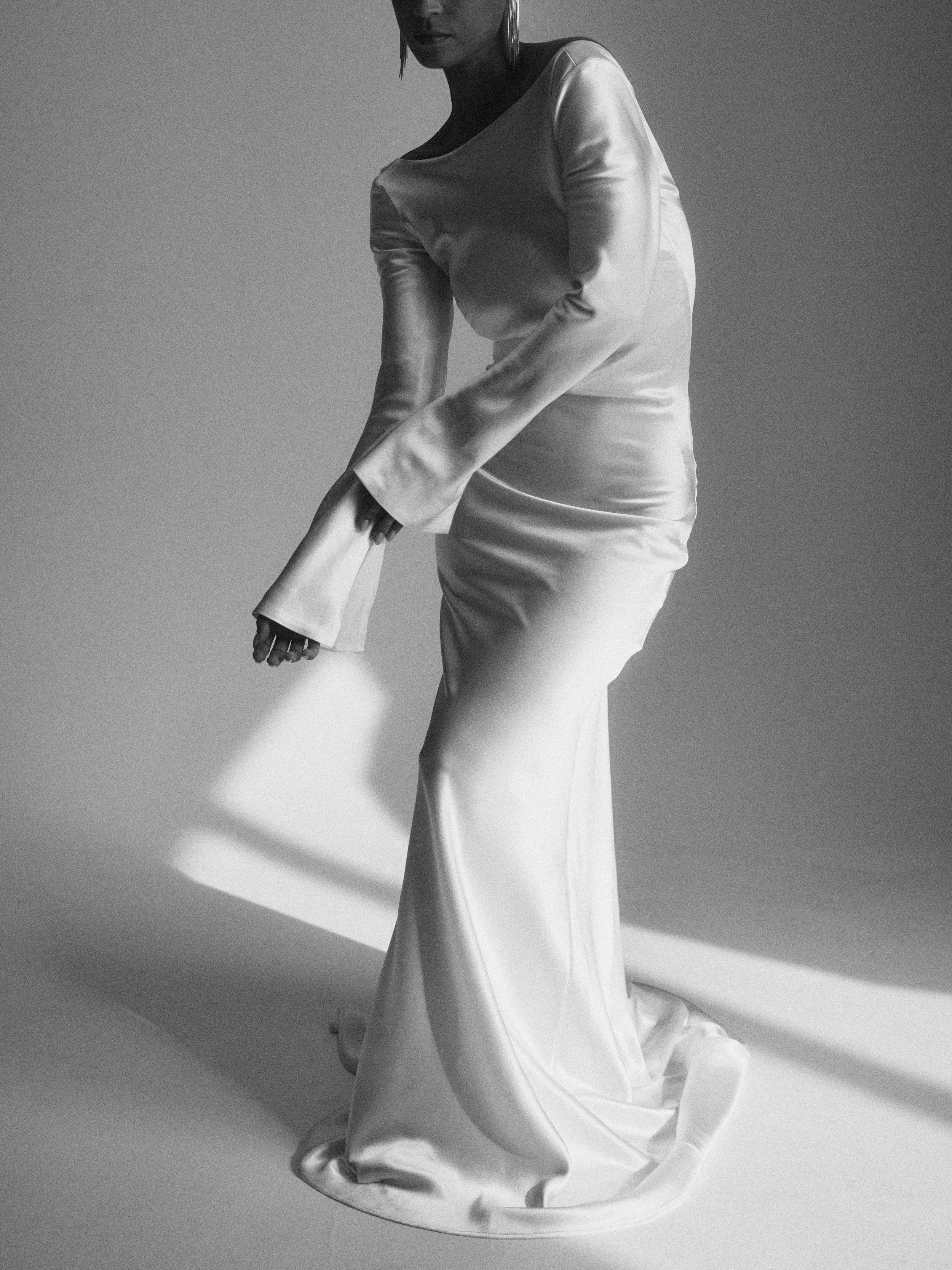 Black and white photo of a woman wearing a long, silky dress, standing with her body slightly bent forward and her right arm extended downwards. The dress has long sleeves and a round neckline, and she is positioned against a plain background.