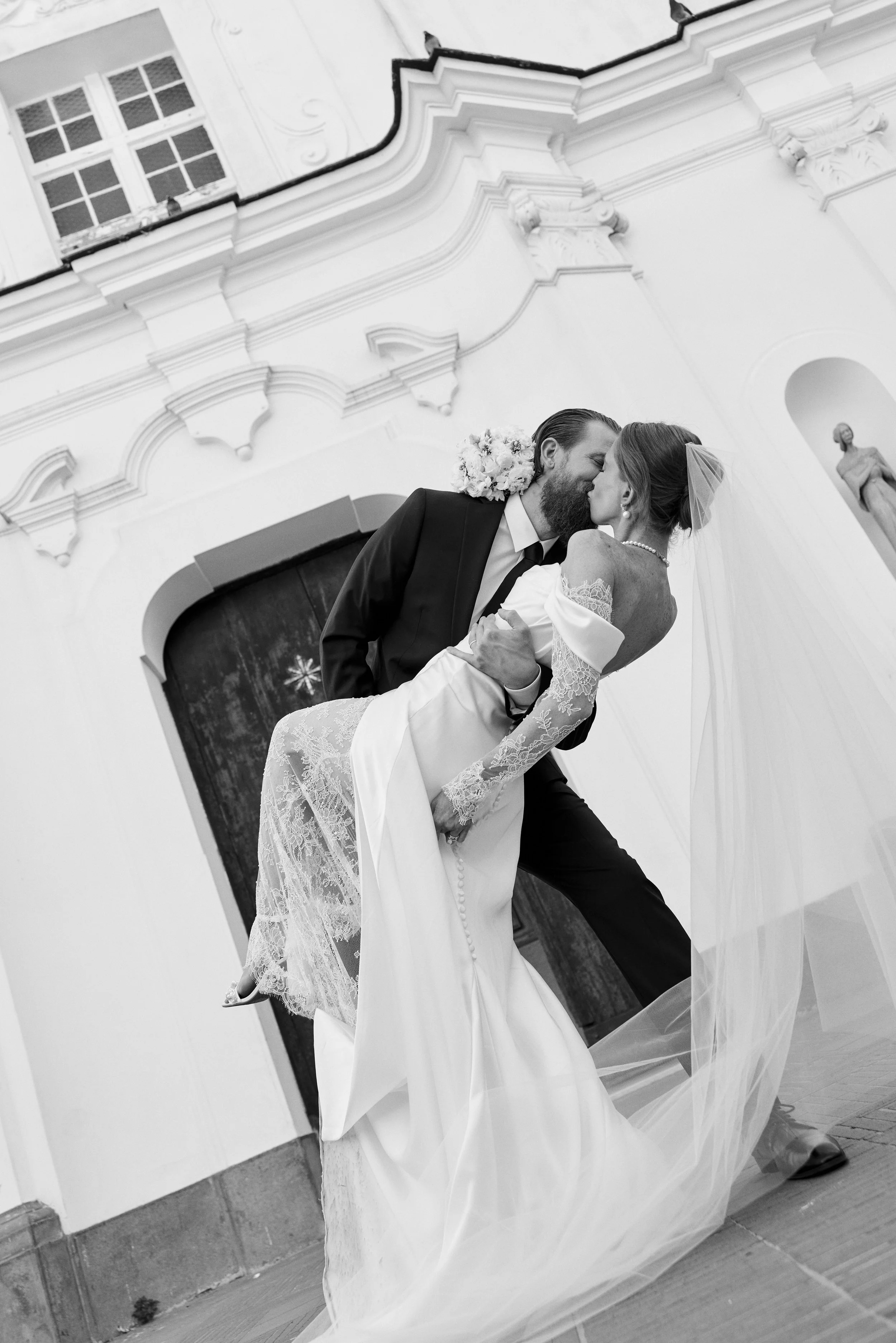 A black and white photo of a groom holding a bride who is leaning back during a wedding ceremony in a decorated church with statues and ornate architectural details.