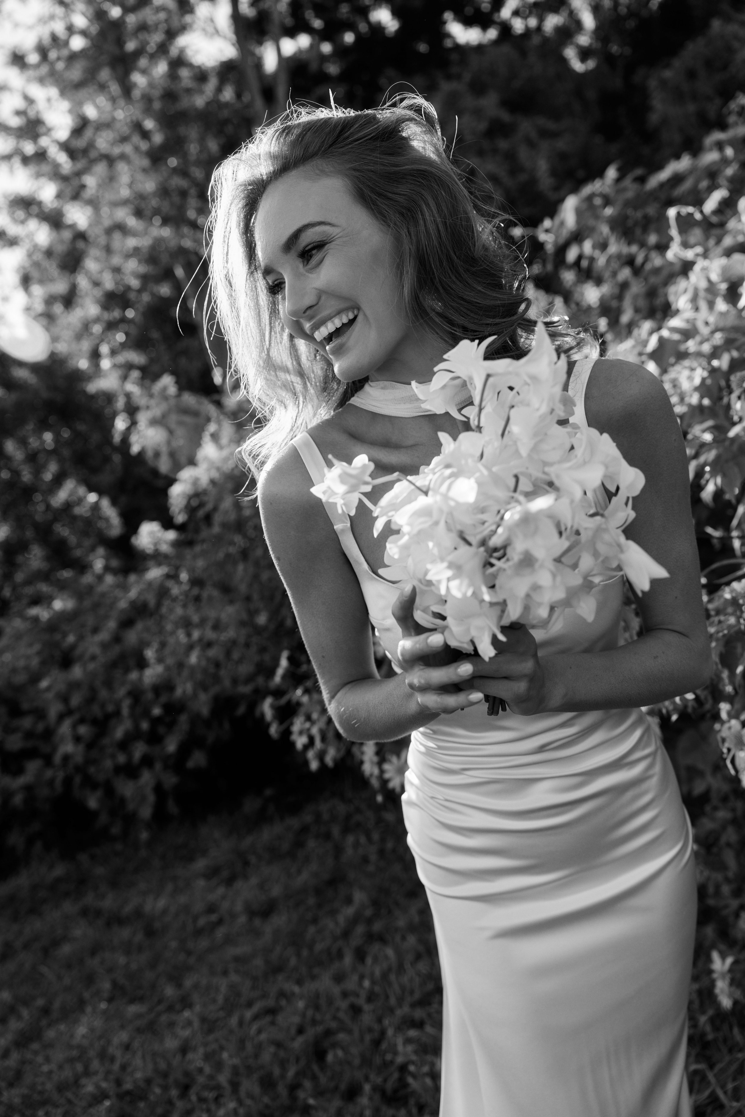 A woman in a light-colored dress holding a bouquet of flowers, smiling outdoors with trees and foliage in the background.