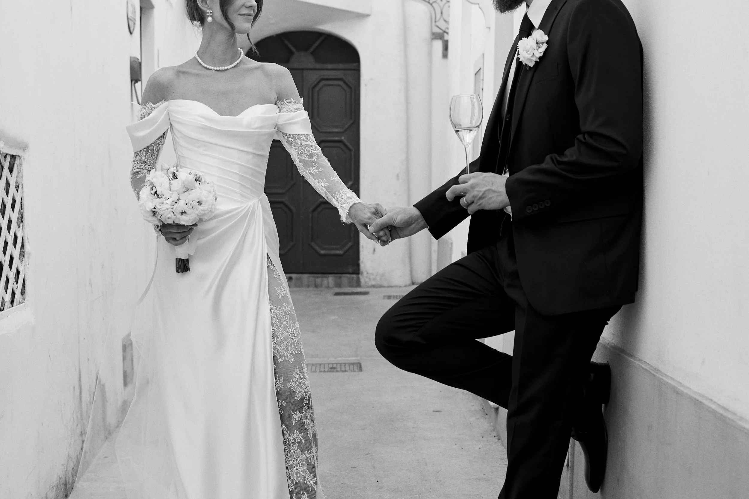 A bride and groom holding hands at a wedding, with the bride wearing a wedding dress and holding a bouquet, and the groom leaning against a wall holding a glass of champagne.