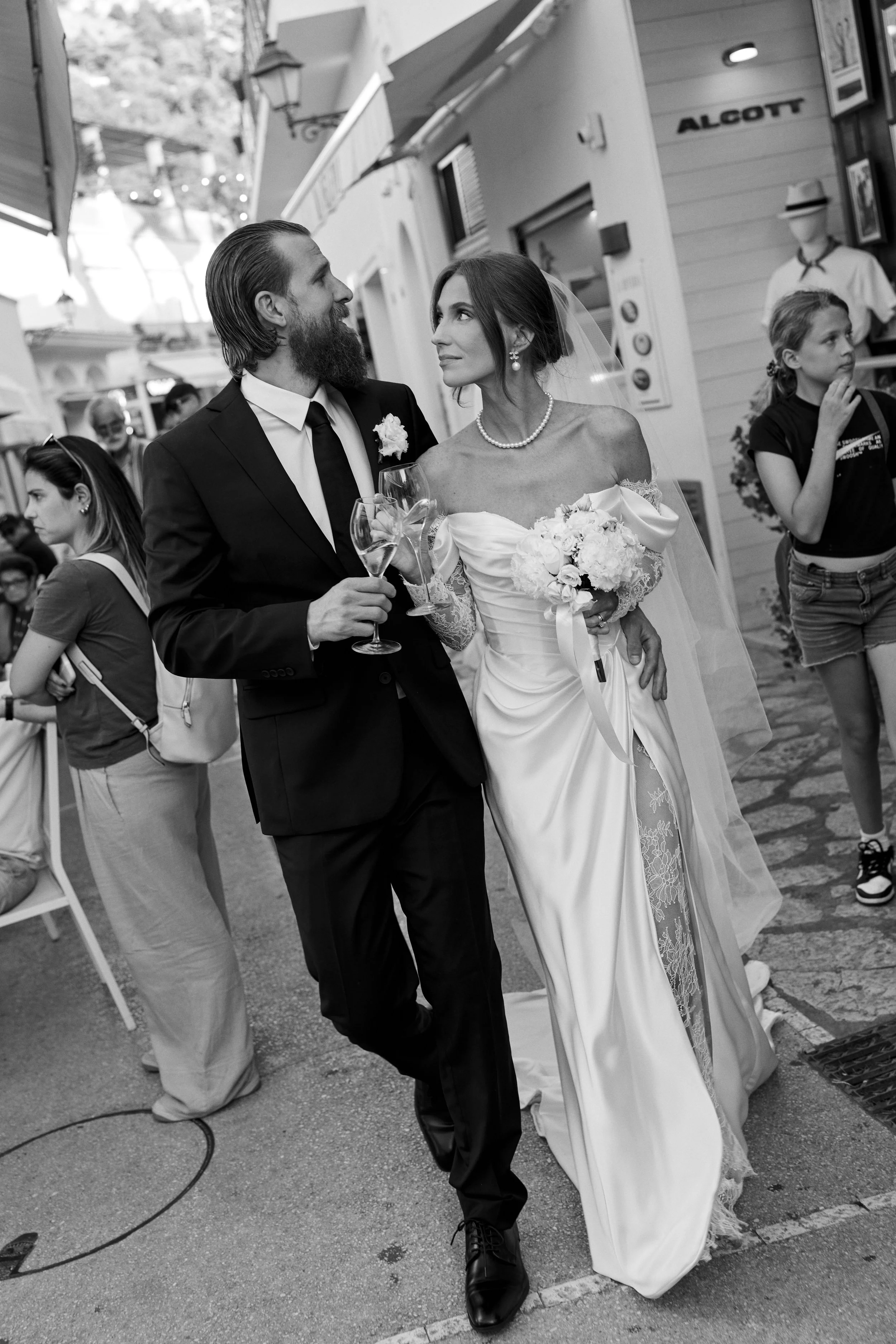 A bride and groom walking together, holding glasses of champagne, at their wedding celebration outdoors.