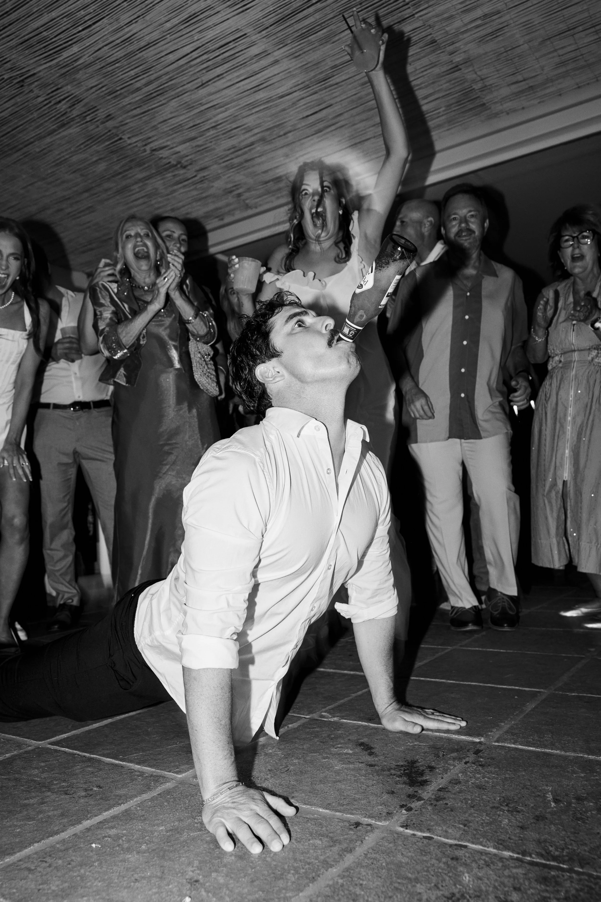 A man performing a handstand on the floor while balancing a bottle on his mouth during a party, surrounded by people cheering and celebrating.
