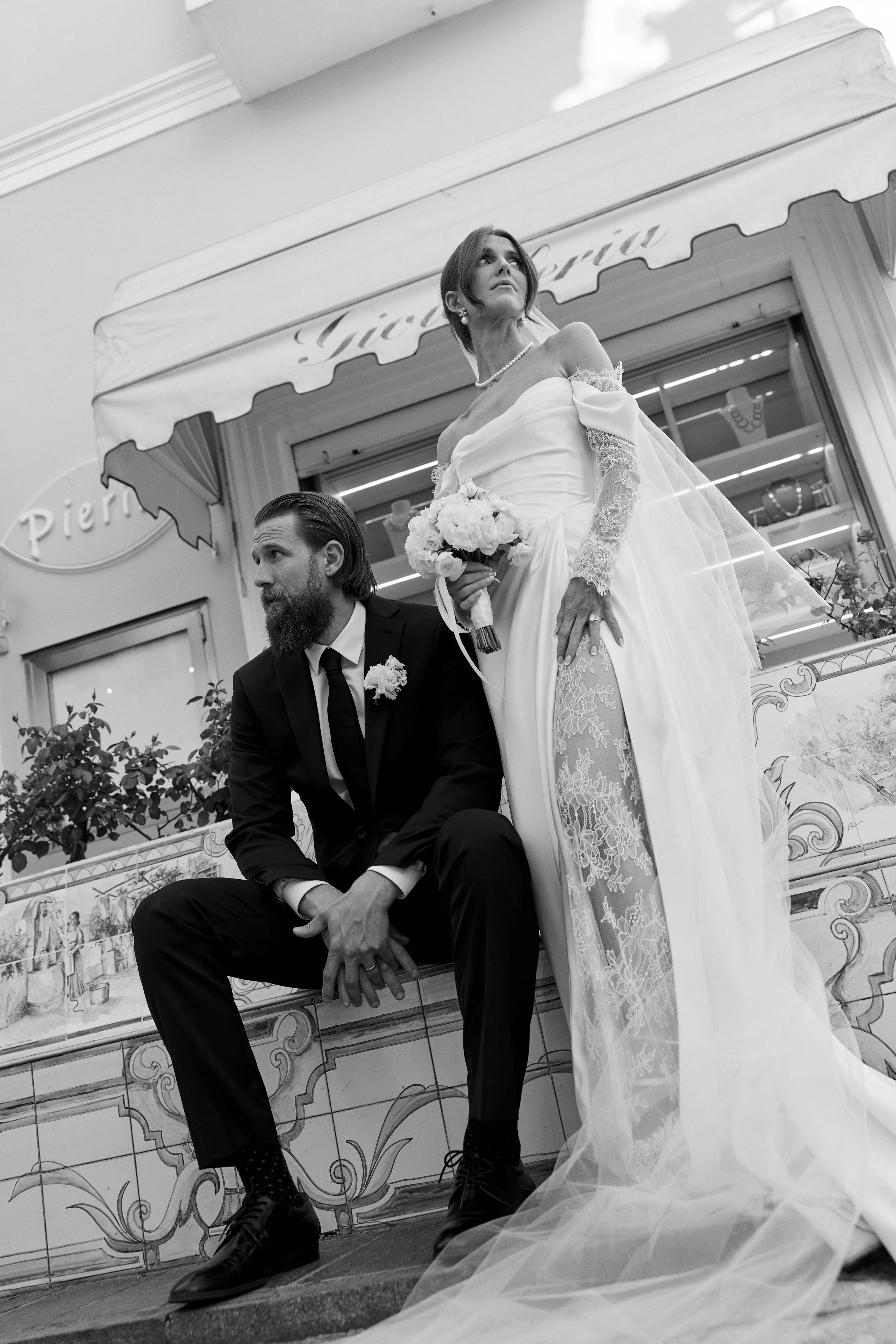 Black and white photo of a bride and groom on their wedding day, with the bride standing and the groom sitting in front of a decorative building with a sign that reads 'Pierina.' The bride is holding a bouquet and wearing a lace wedding gown with off