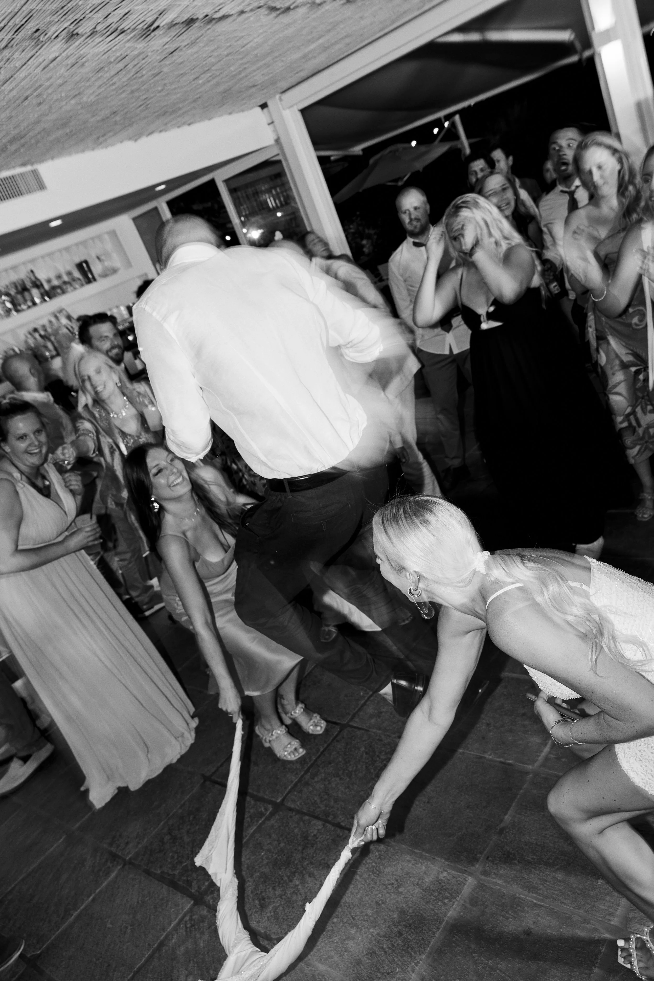 A lively party scene with people dancing and having fun indoors, some watching as two women and a man participate in a game involving pulling a rope. The atmosphere appears energetic and joyful.