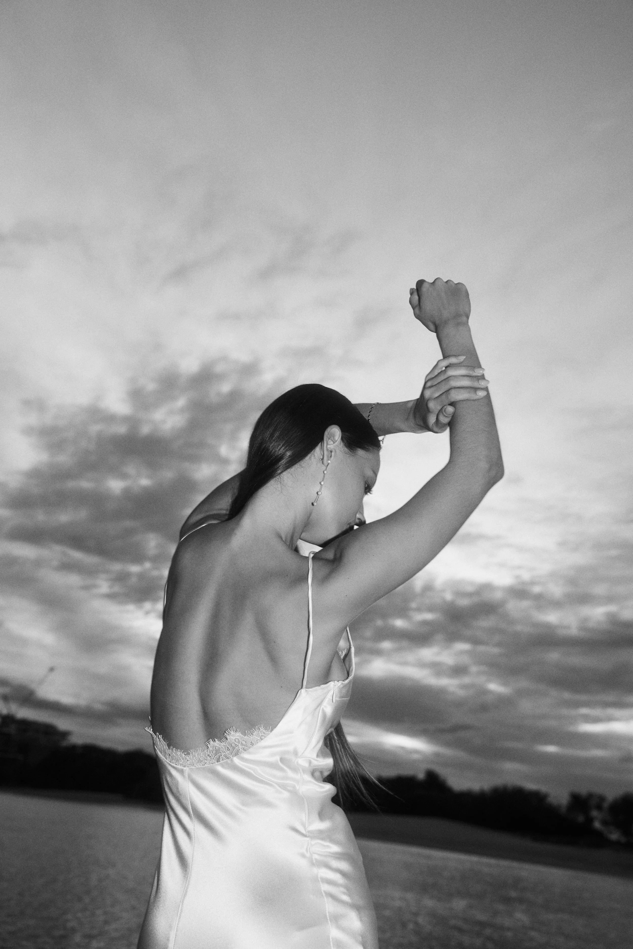 A woman in a satin slip dress is standing in an outdoor setting at sunset. She has long hair, is wearing a dangling earring, and is holding her arms up in a flexed pose.