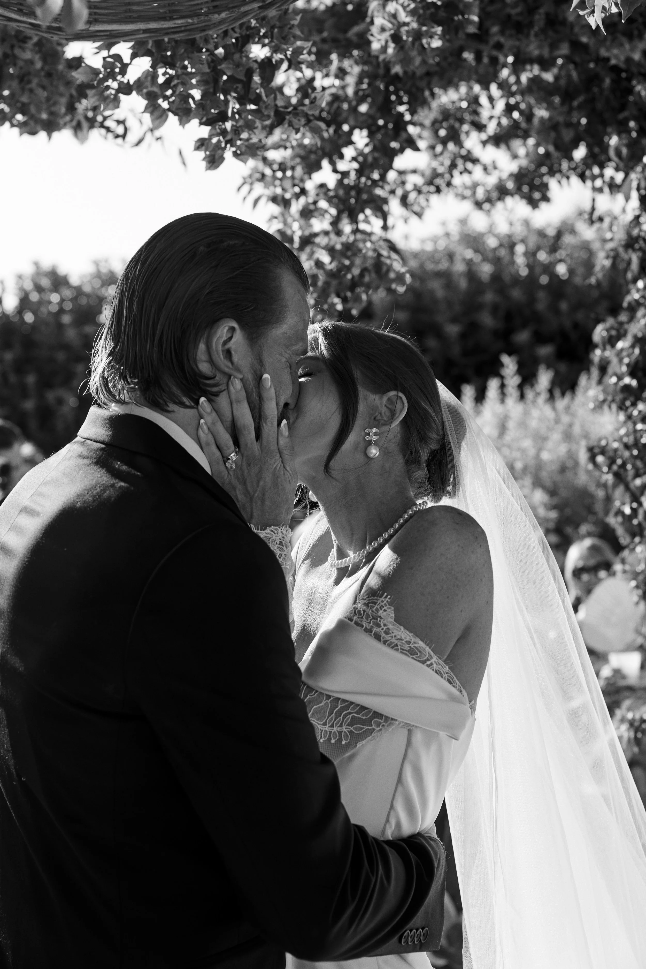 Black and white photo of a bride and groom sharing a kiss outdoors on their wedding day, with sunlight filtering through trees in the background.