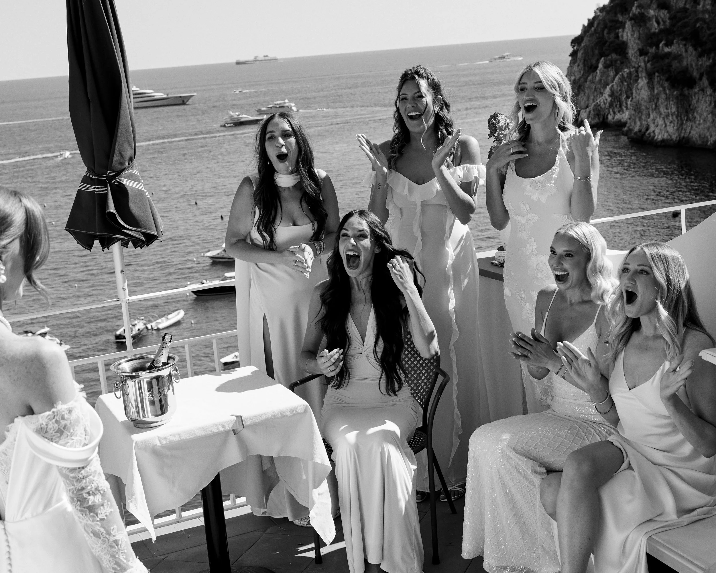 Group of women in wedding dresses celebrating on a balcony overlooking the ocean, with boats in the background.