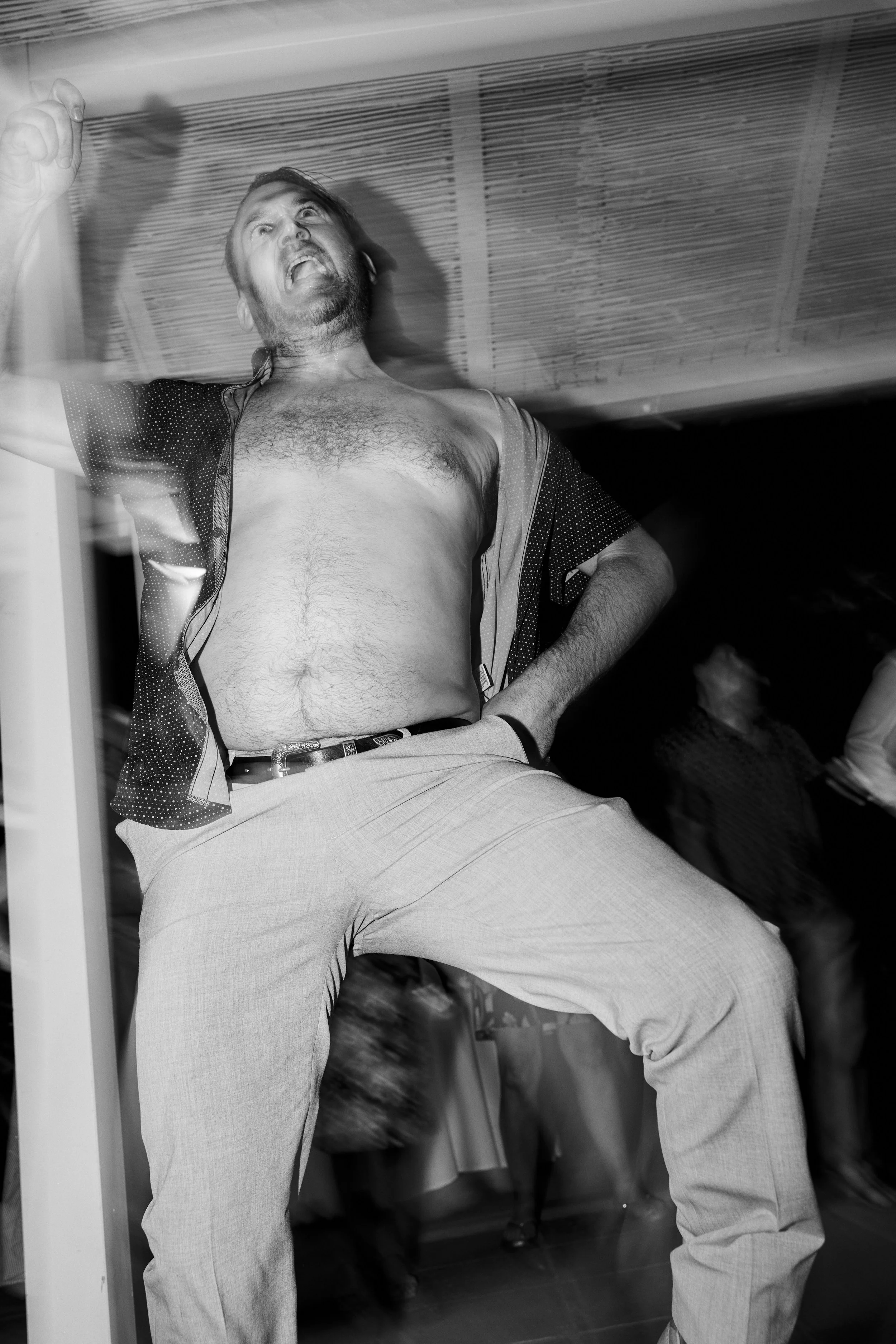 A man with a beard, shirt off, standing with hands on his hips, looking upward with an expressive face, in a room with a low ceiling.