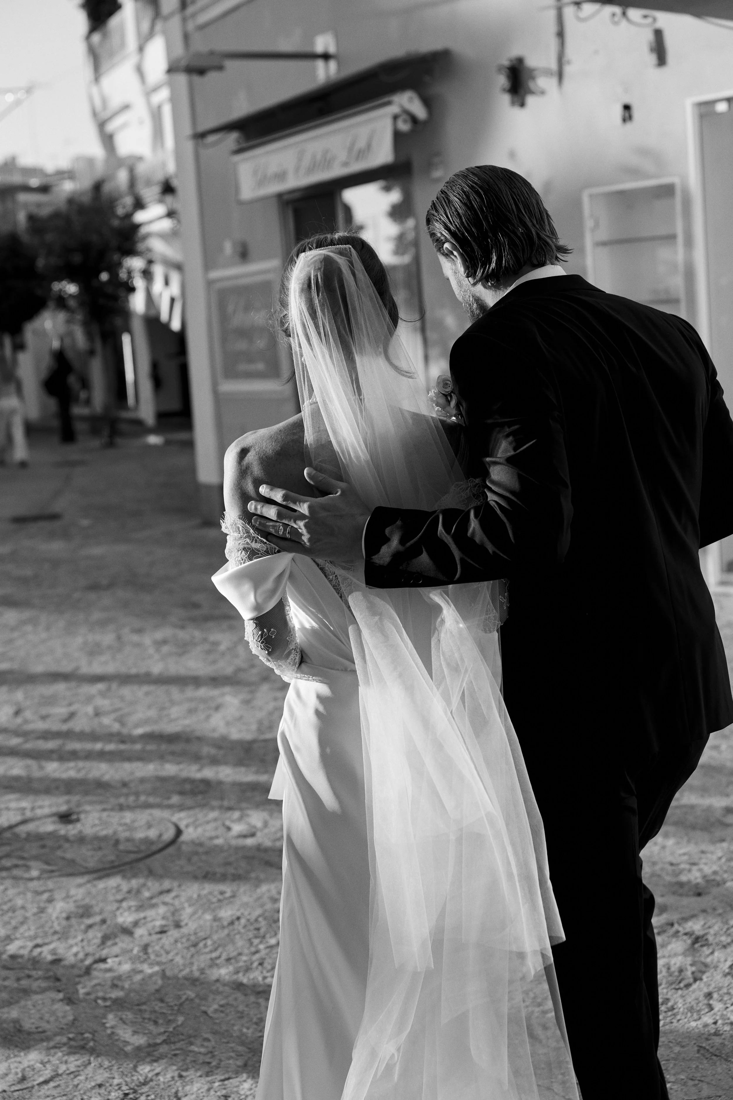 A black-and-white photo of a bride in a wedding dress and veil with a man, possibly her groom, standing close after a wedding ceremony outdoors.