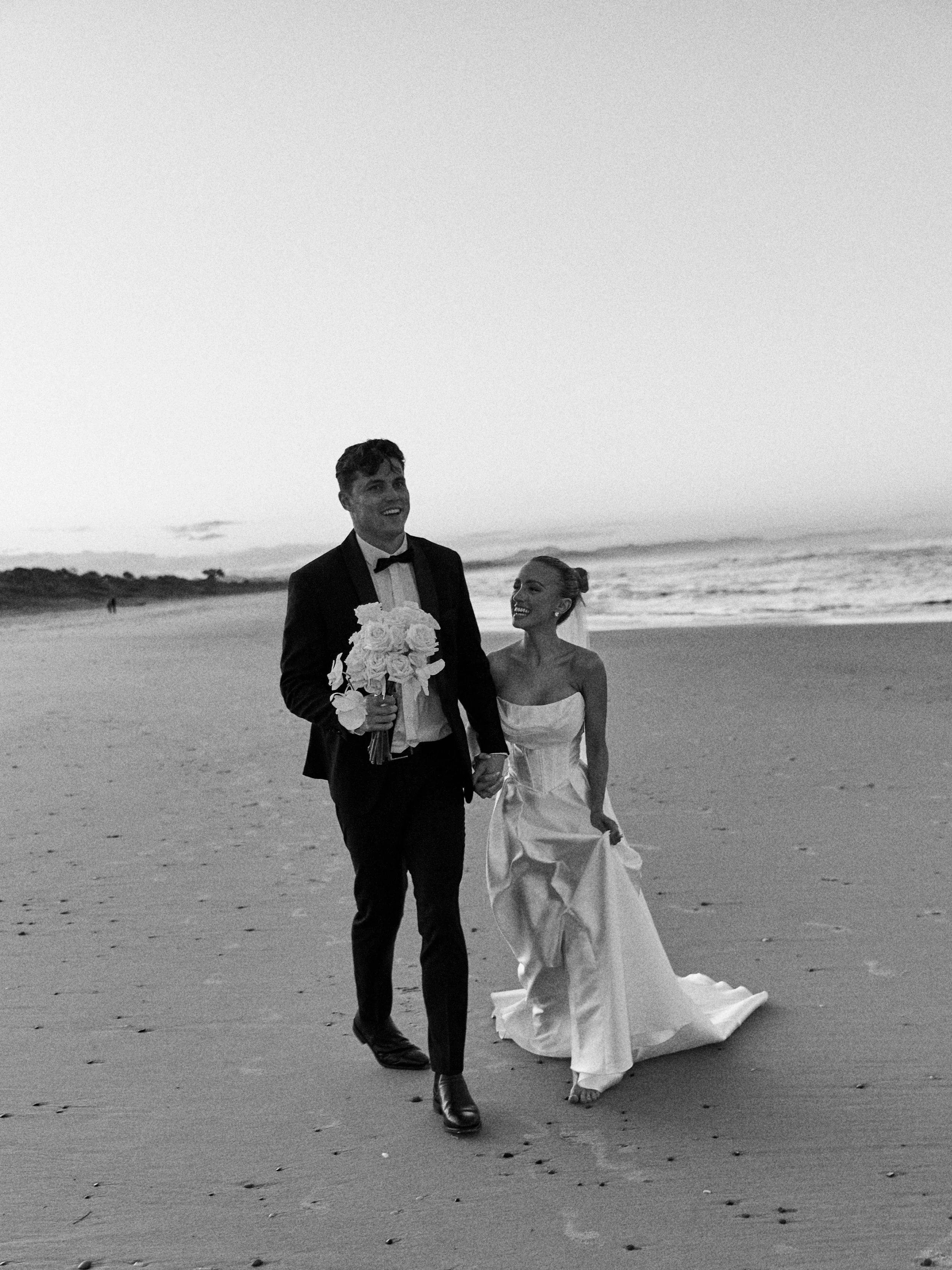 A newlywed couple walking on the beach at sunset, smiling and holding hands. The groom is holding a bouquet of flowers. The scene is in black and white.