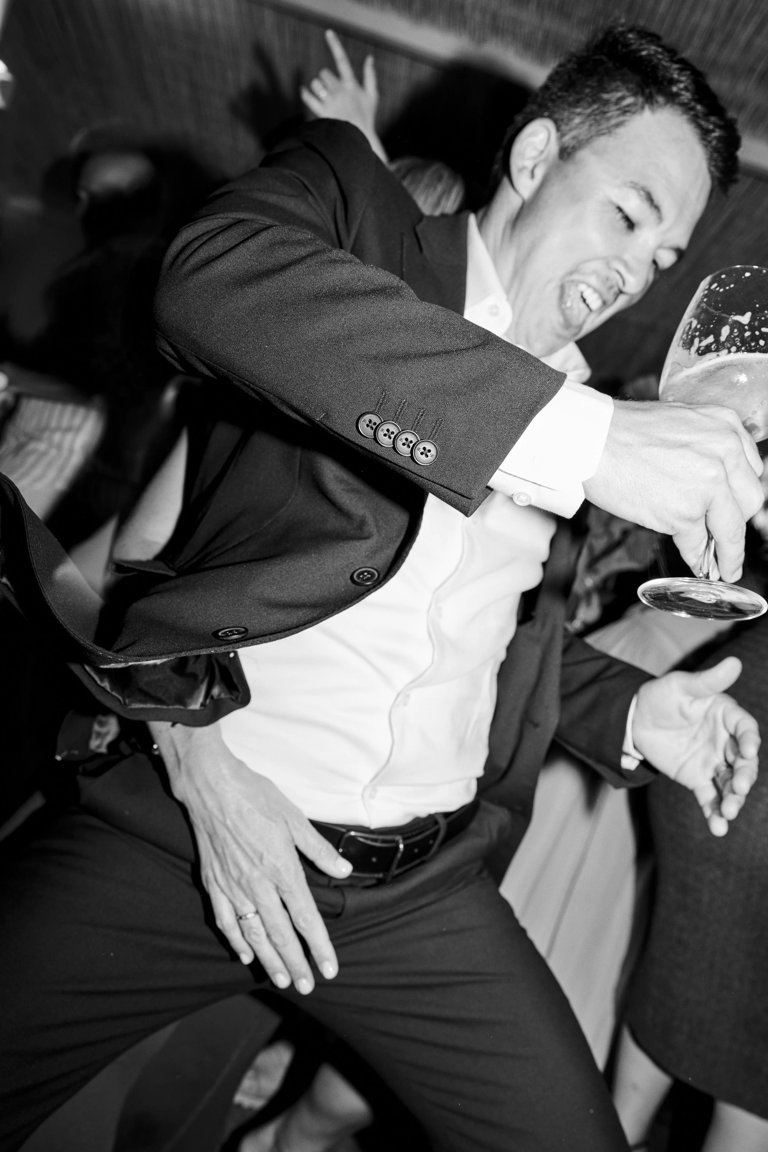 A man in a suit and white shirt is laughing and reaching for a glass of beer, sitting at a table in a social setting.
