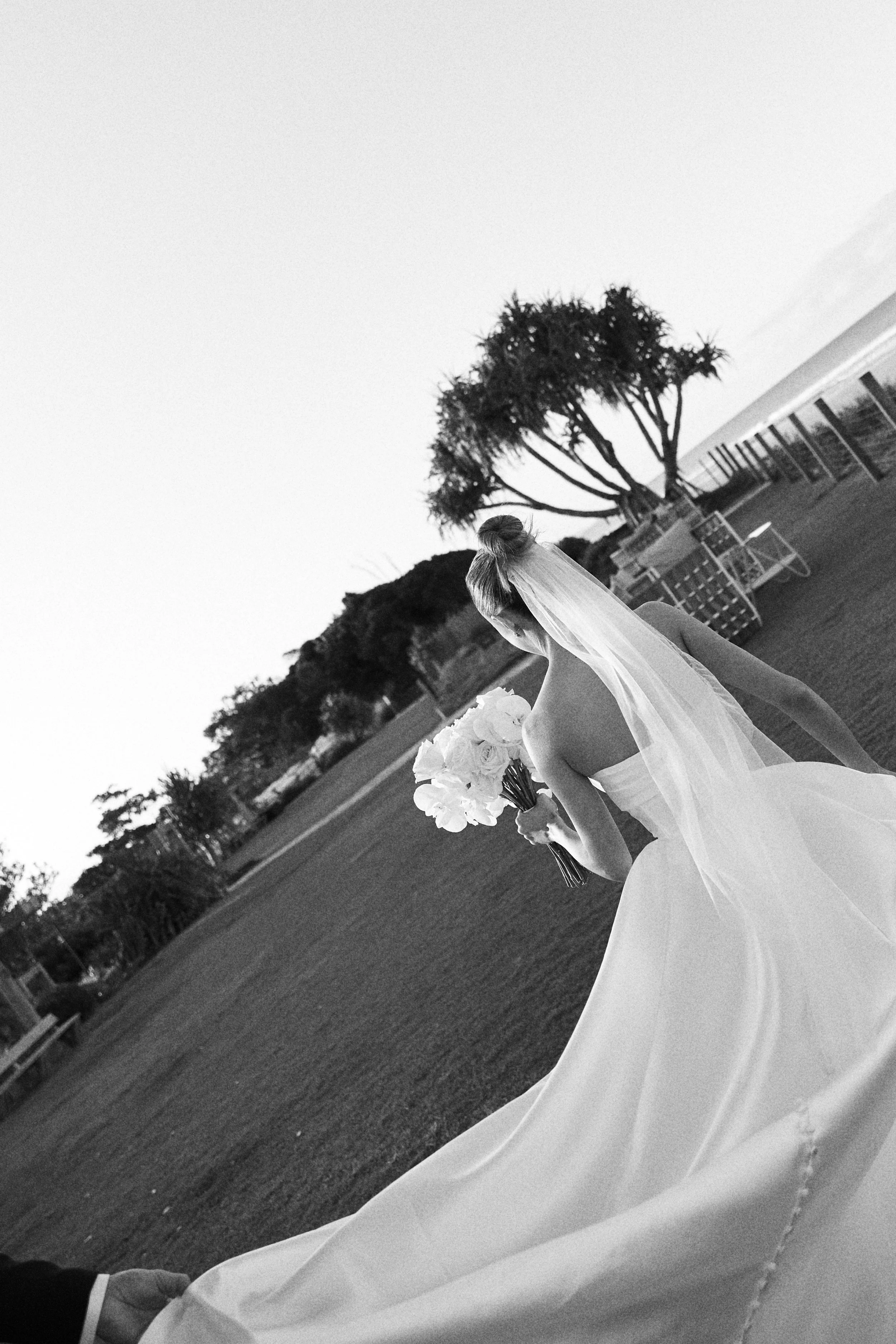 Black and white photo of a bride holding a bouquet of flowers on a grassy outdoor area near the ocean.