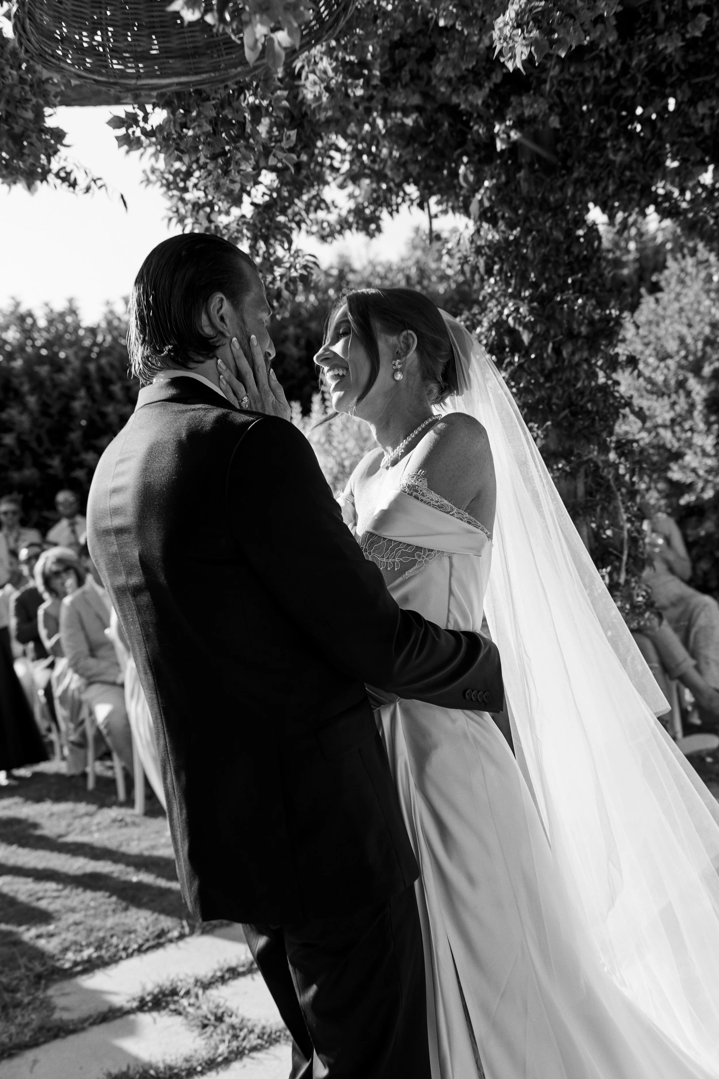 A black and white photo of a bride and groom at their wedding, smiling and touching faces, outdoors under trees with guests in the background.