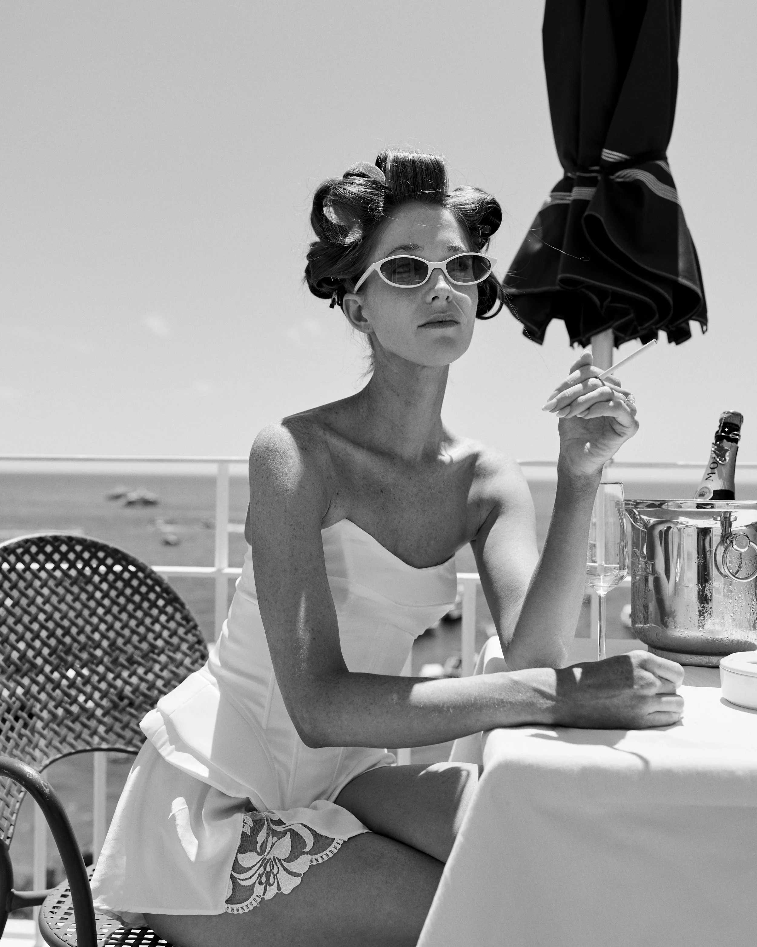 A woman wearing sunglasses and a strapless dress sitting at a table with a beach view behind her, with an umbrella and a bottle in an ice bucket.