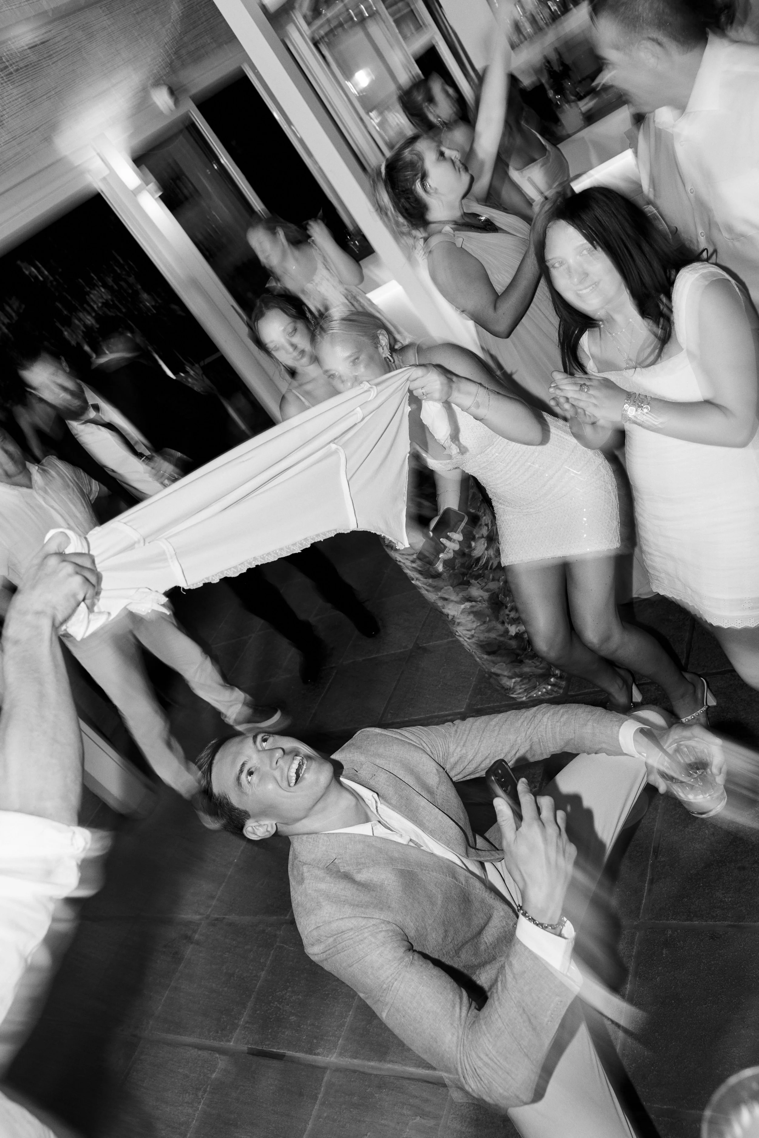 People celebrating and playing games at a party, with some pulling a long cloth and others smiling and clapping, in black and white.