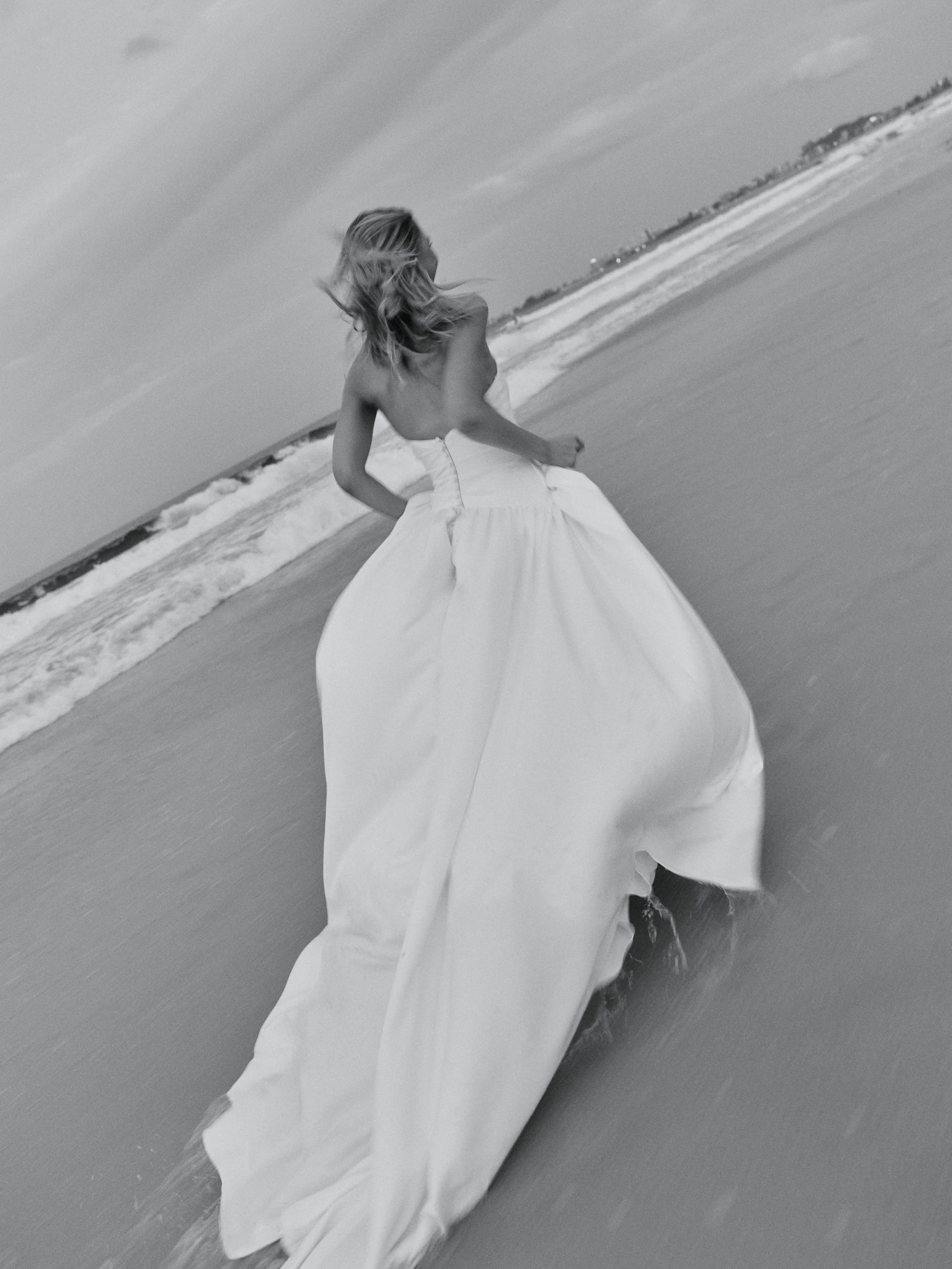 A woman in a strapless wedding dress walking along the beach at or near sunset in black and white.
