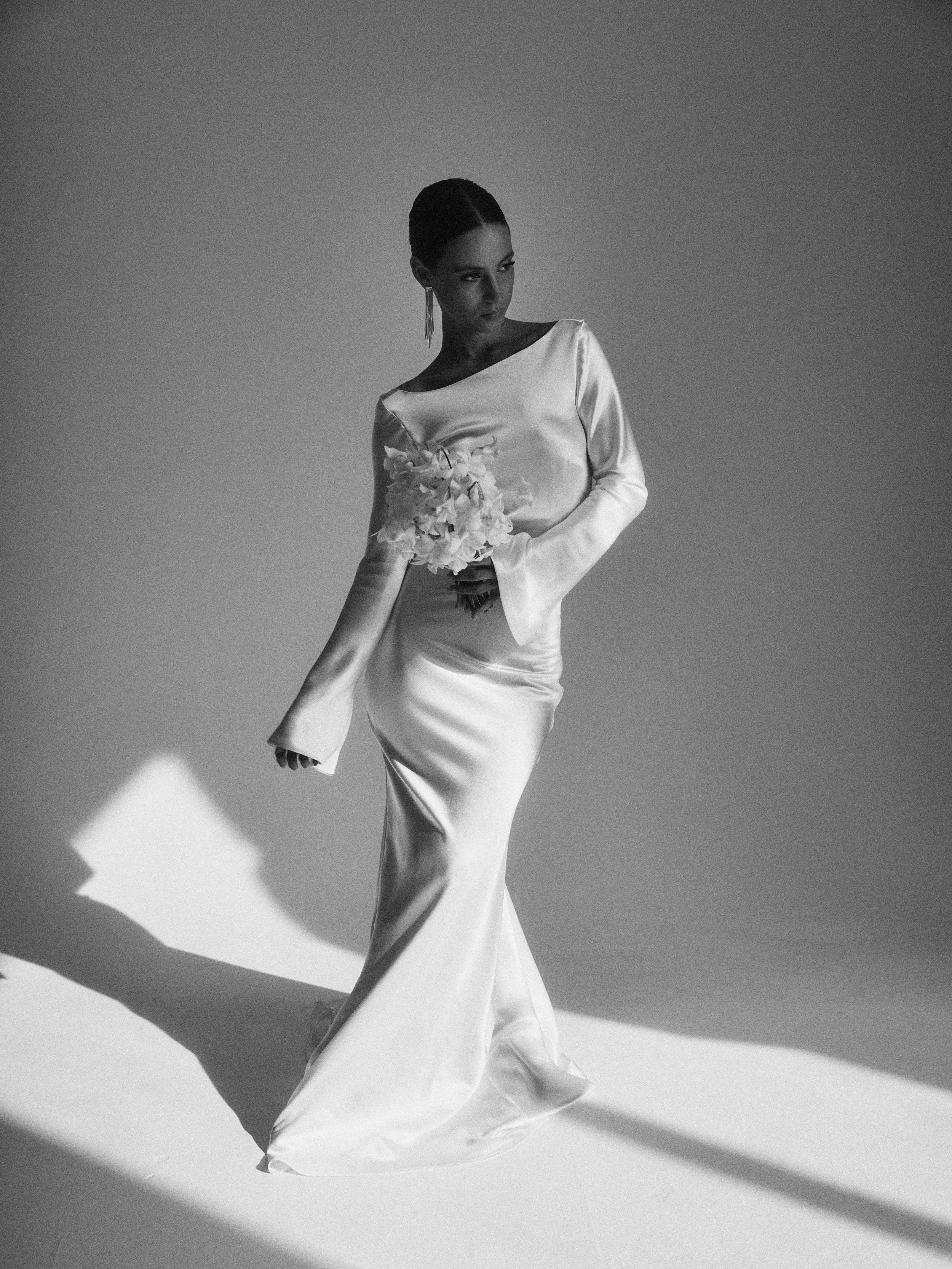 A woman in a long satin dress holding a bouquet of flowers, standing against a plain background with shadows cast on the floor.