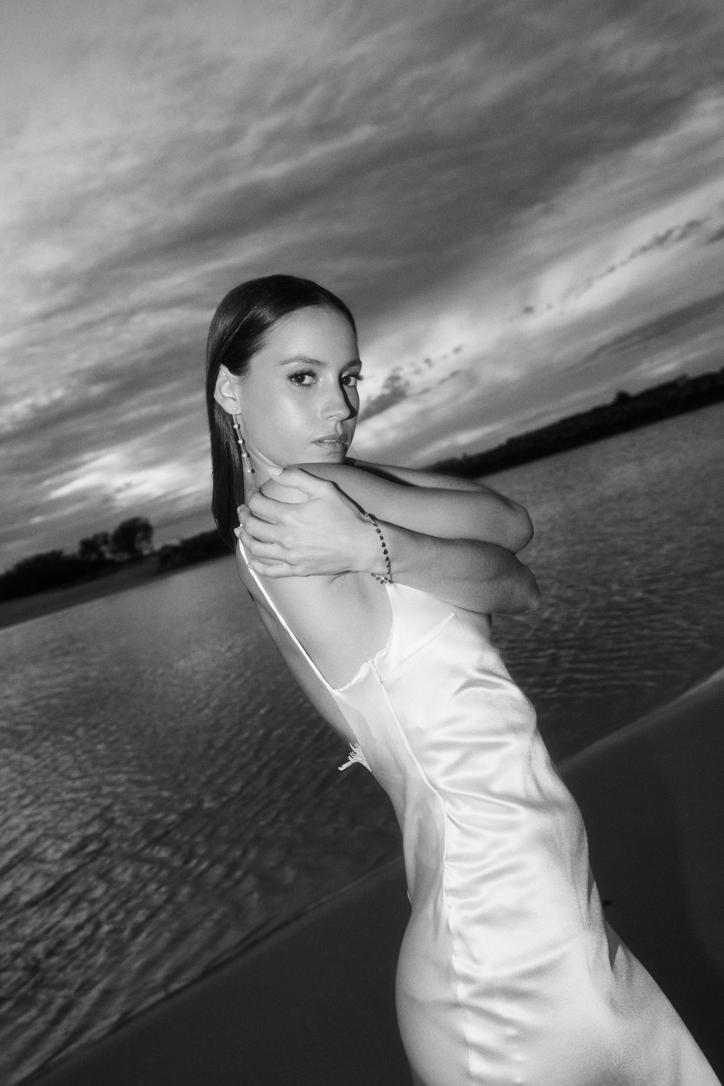 A woman in a satin dress posing by water with a cloudy sky in black and white.