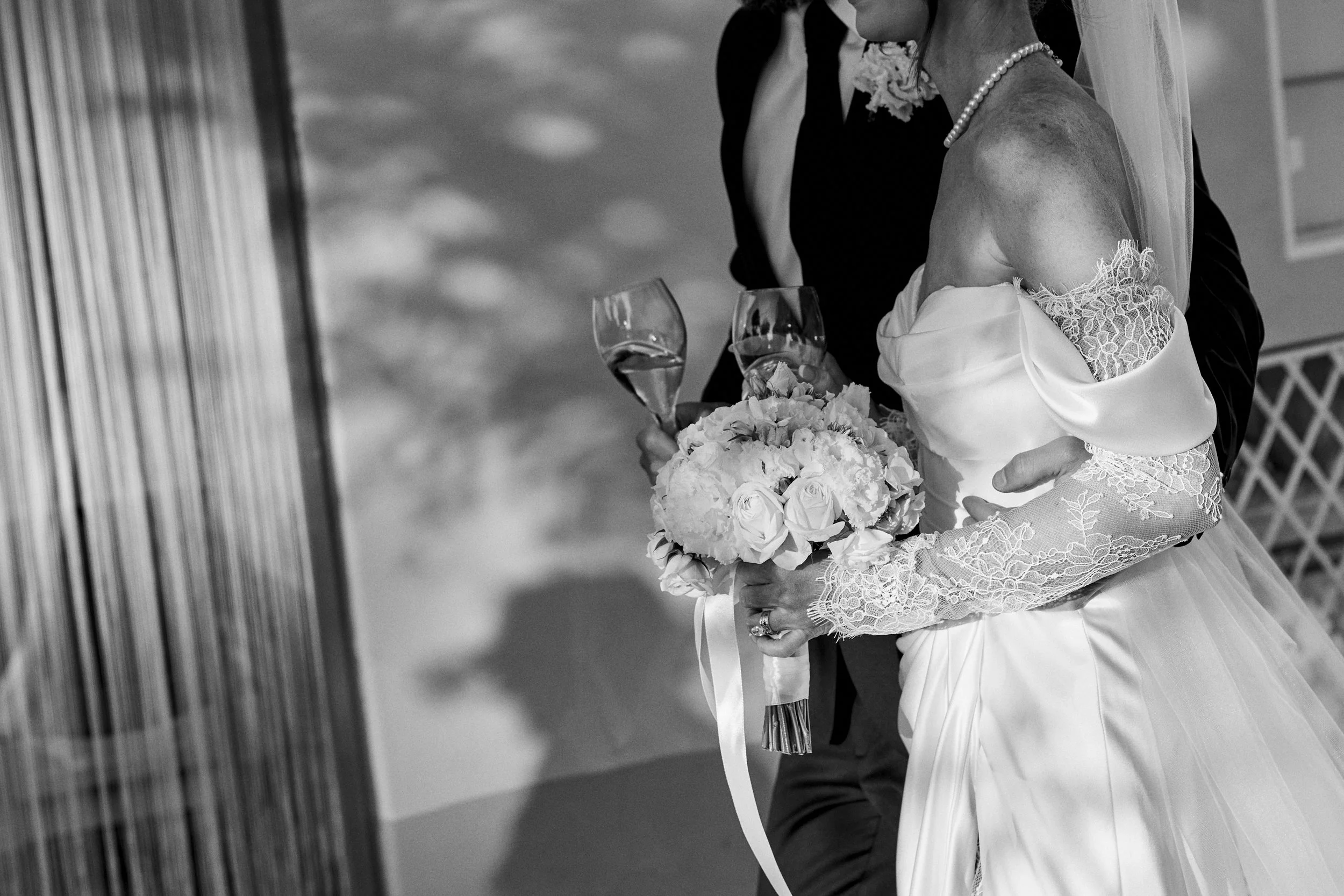 Black and white photograph of a bride and groom holding champagne glasses, with the bride holding a bouquet of roses and hydrangeas, and wearing an off-shoulder wedding gown with lace sleeves.