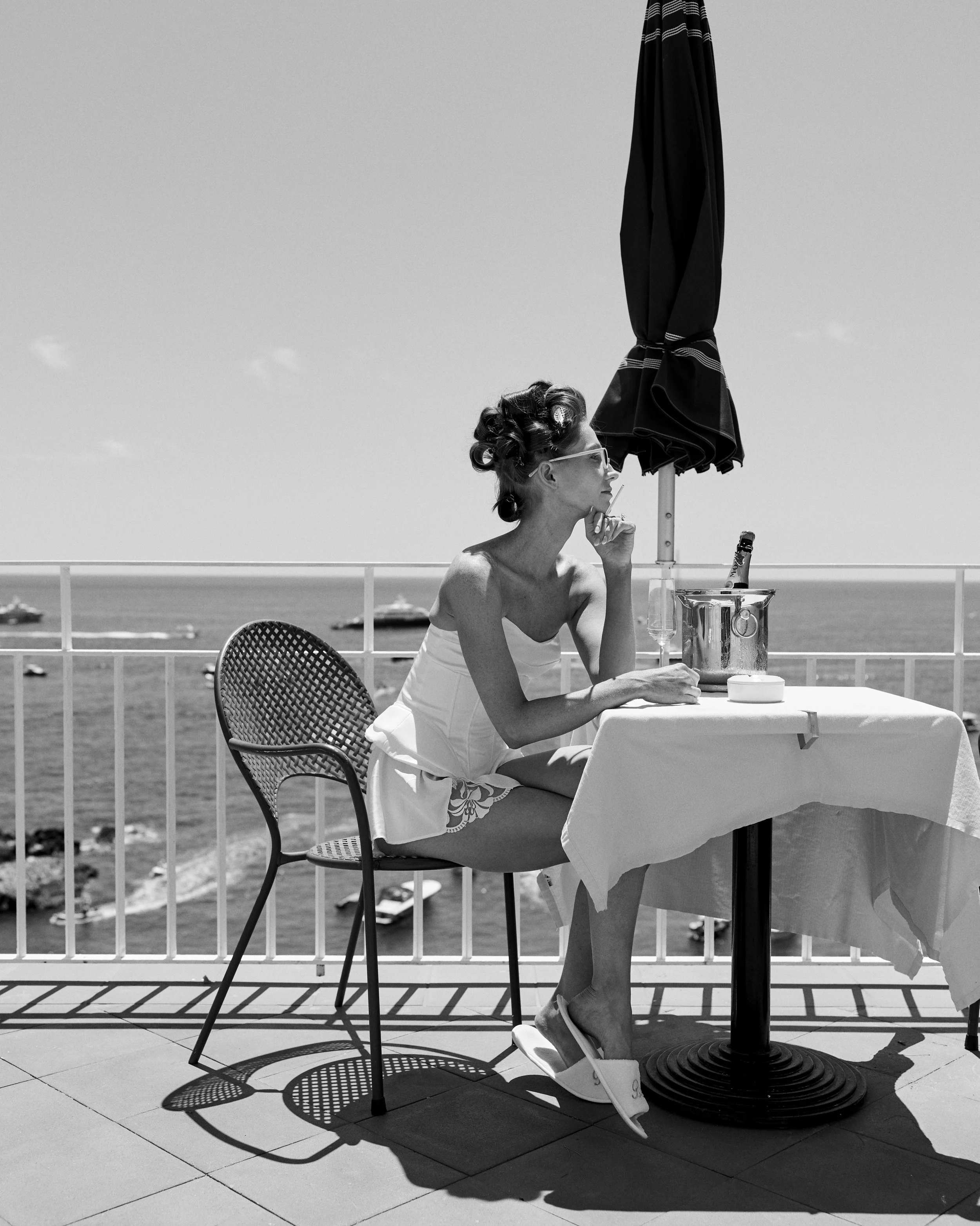 A woman sitting at a table on a balcony overlooking the ocean, wearing sunglasses, a strapless top, shorts, and slippers, with an umbrella providing shade.