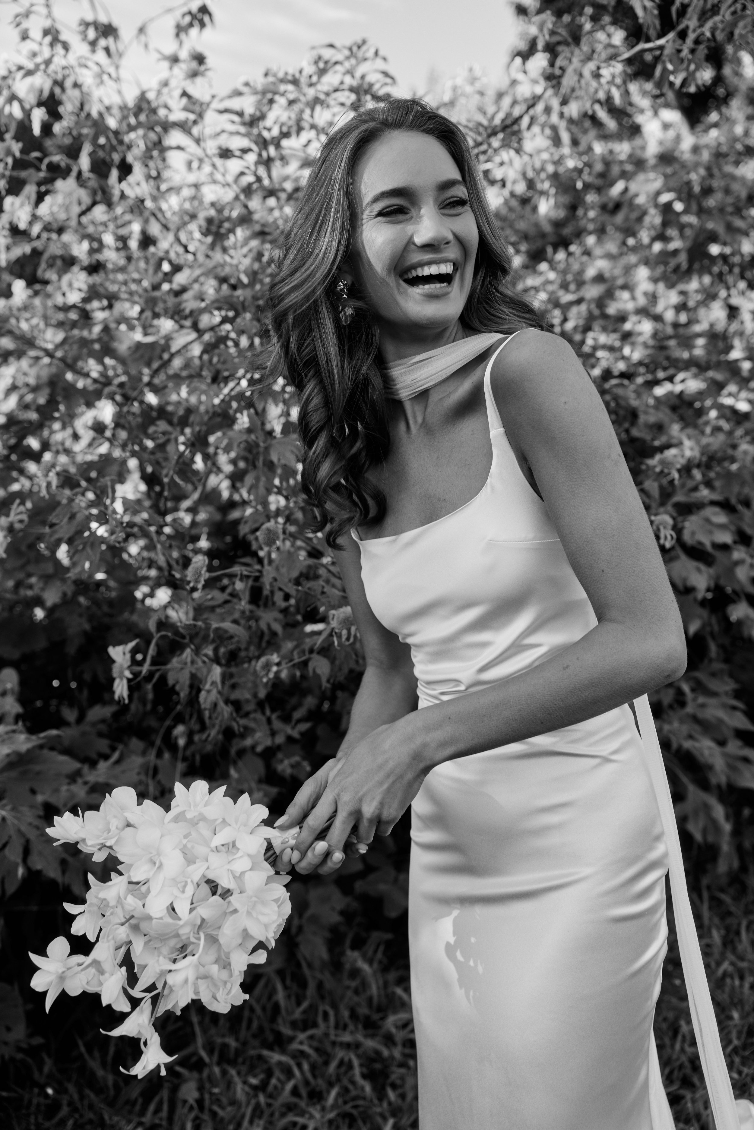 A woman in a wedding dress holding a bouquet of flowers, smiling and laughing outdoors surrounded by foliage.