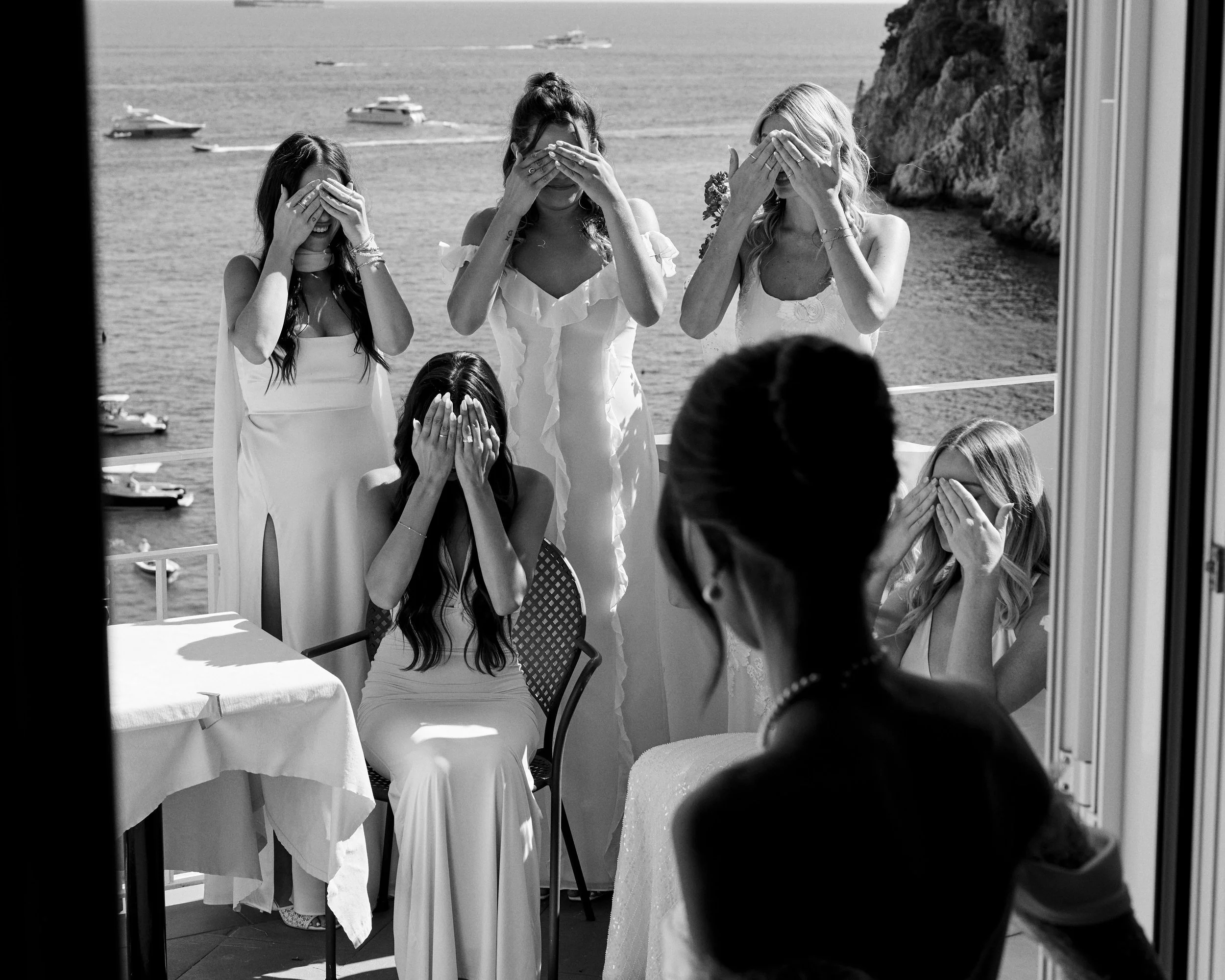 A group of women in white dresses standing and sitting outside on a balcony, covering their eyes with their hands, with the sea and boats in the background, viewed through a window.