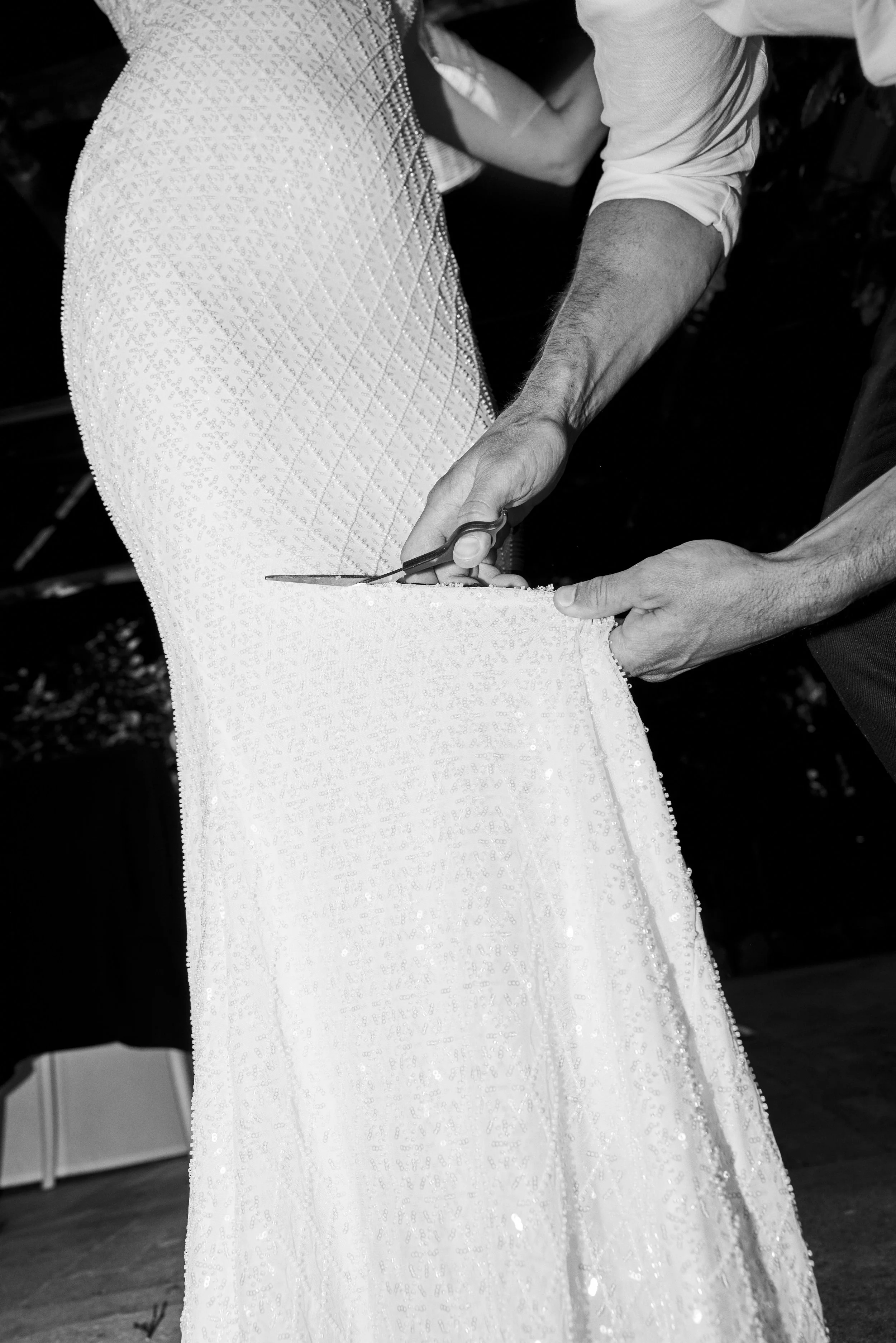 A person in a white dress is being altered or tailored with scissors, possibly at a bridal or formal dress fitting.