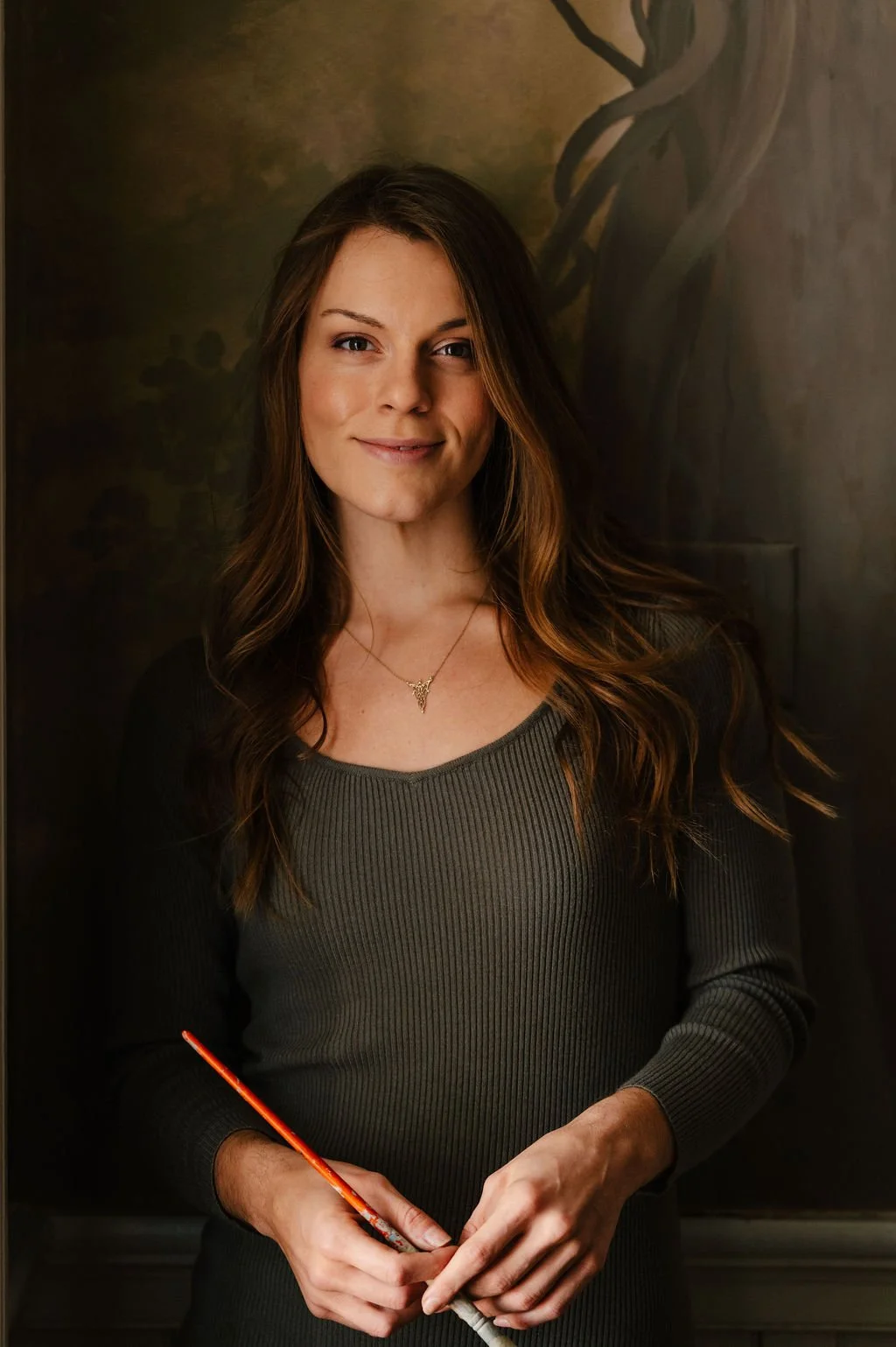 Female professional artist Gabrielle Hasenstab with long wavy brown hair wearing a green ribbed dress and an evanstar pendant necklace standing in front of a dark forest mural.